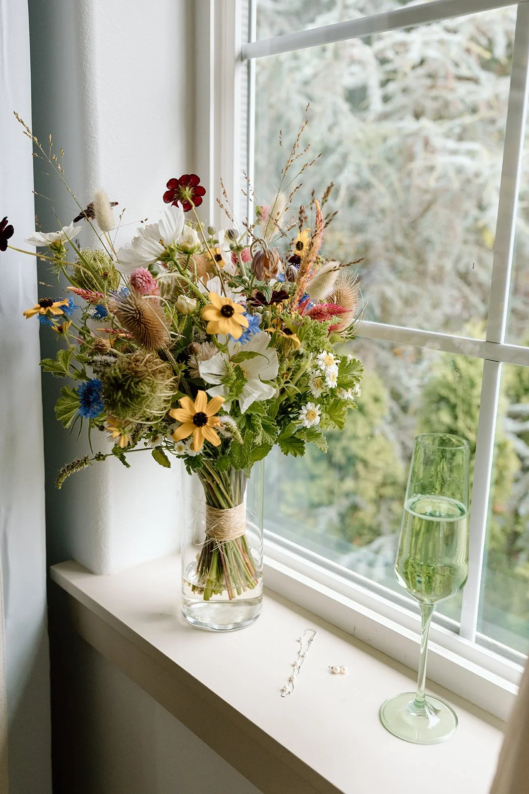 A bouquet of mixed wildflowers in a glass vase on a windowsill, next to a glass of green-colored drink and jewelry, with a view of trees outside.