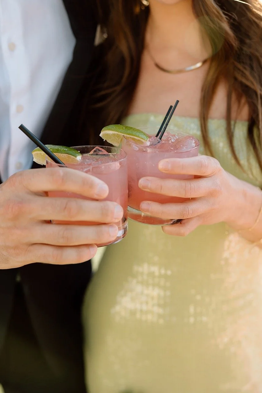 Two people holding pink cocktails garnished with lime wedges and ice, with black straws.