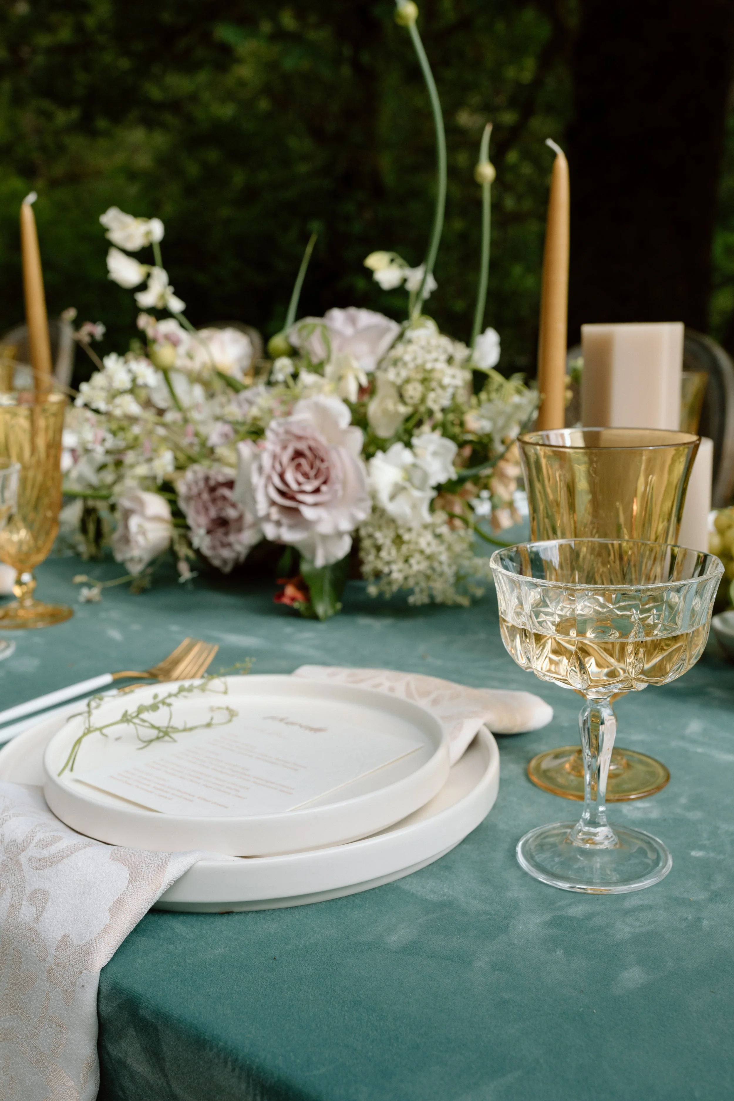 Elegant outdoor table setting with white plates, gold and crystal glassware, and a floral centerpiece with white and blush flowers, candles, and greenery.