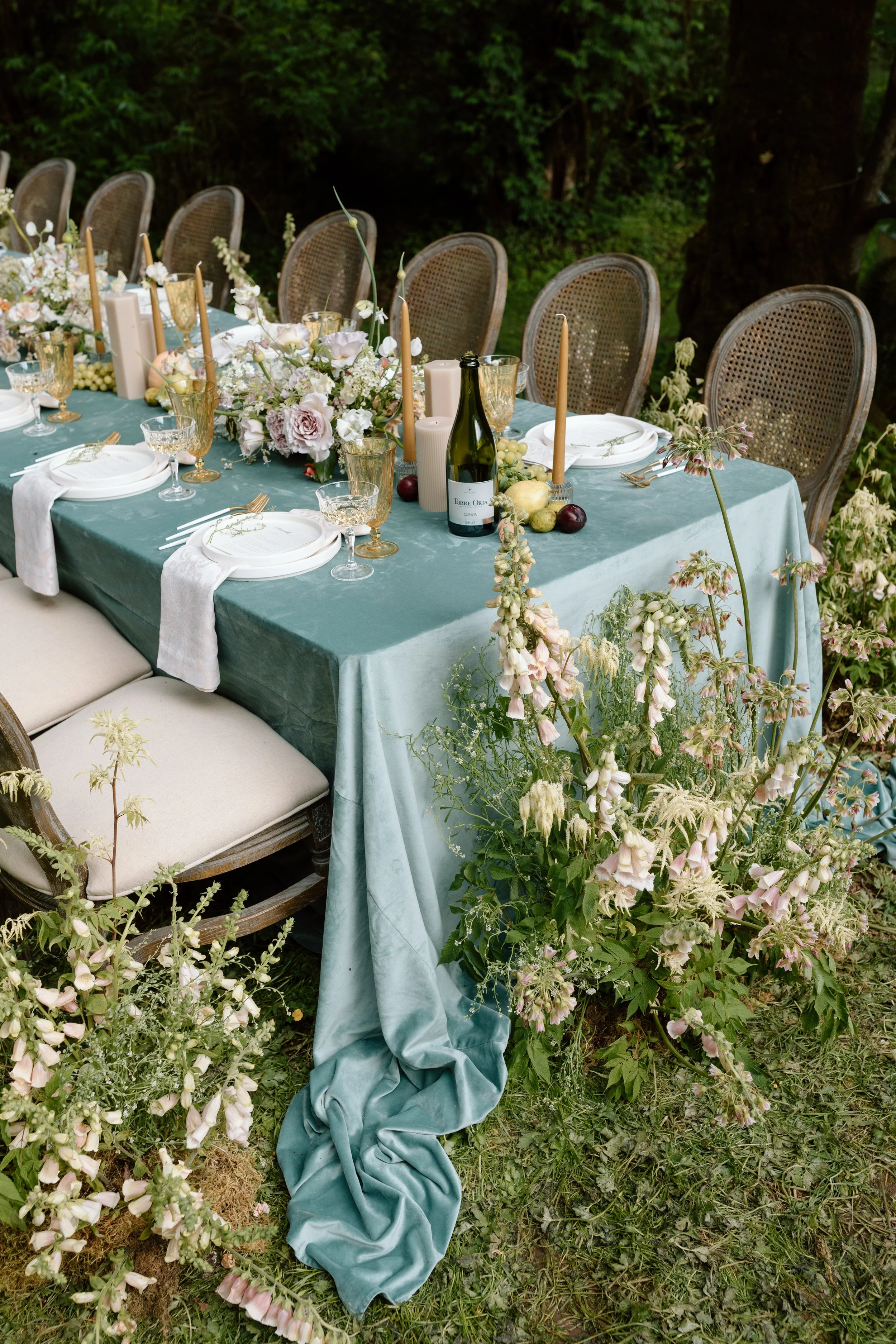 Outdoor dining table set with teal tablecloth, surrounded by rattan chairs, decorated with floral arrangements, candles, and glassware, in a lush garden.