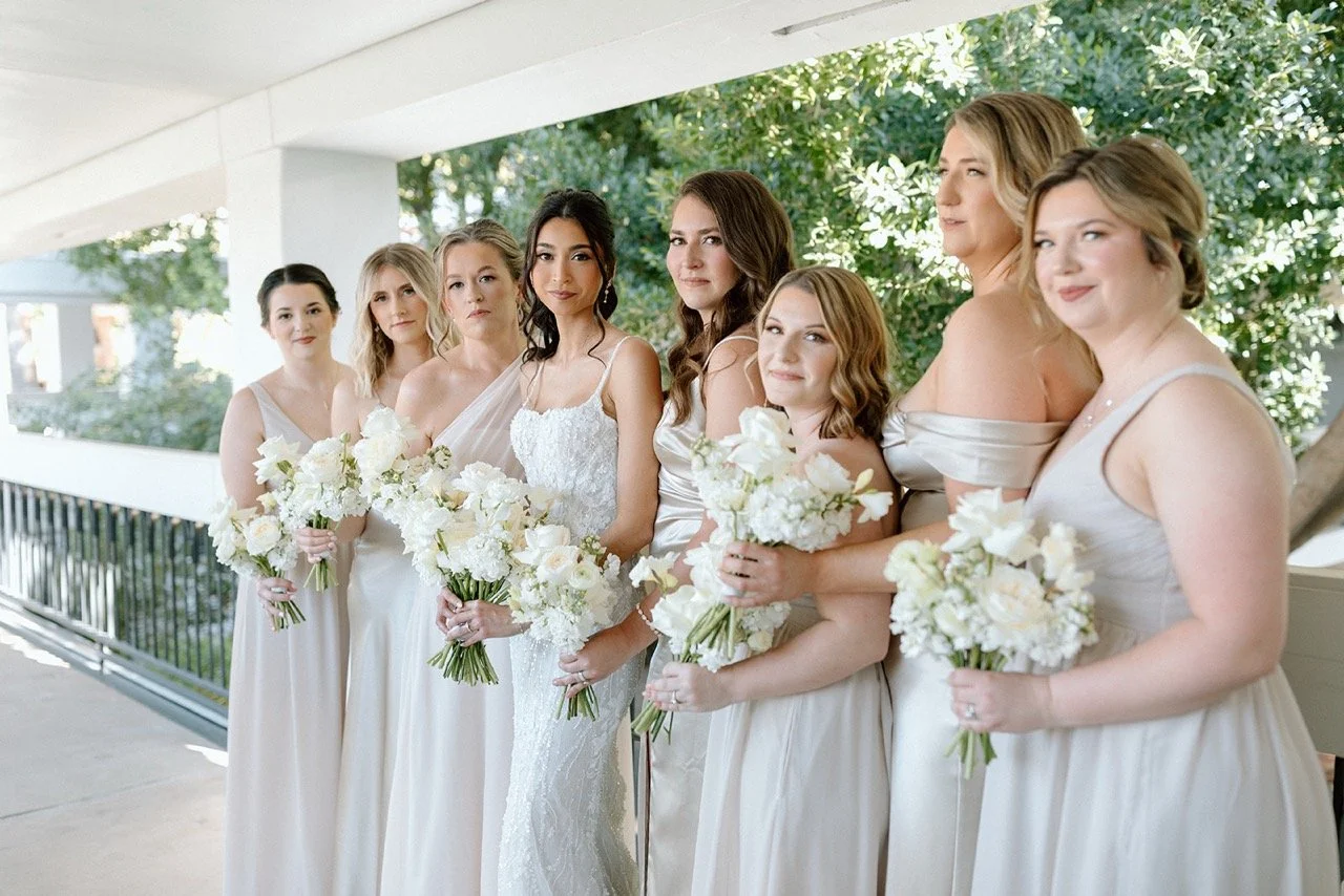Group of eight women in cream dresses holding white bouquets on a veranda with green trees in the background.