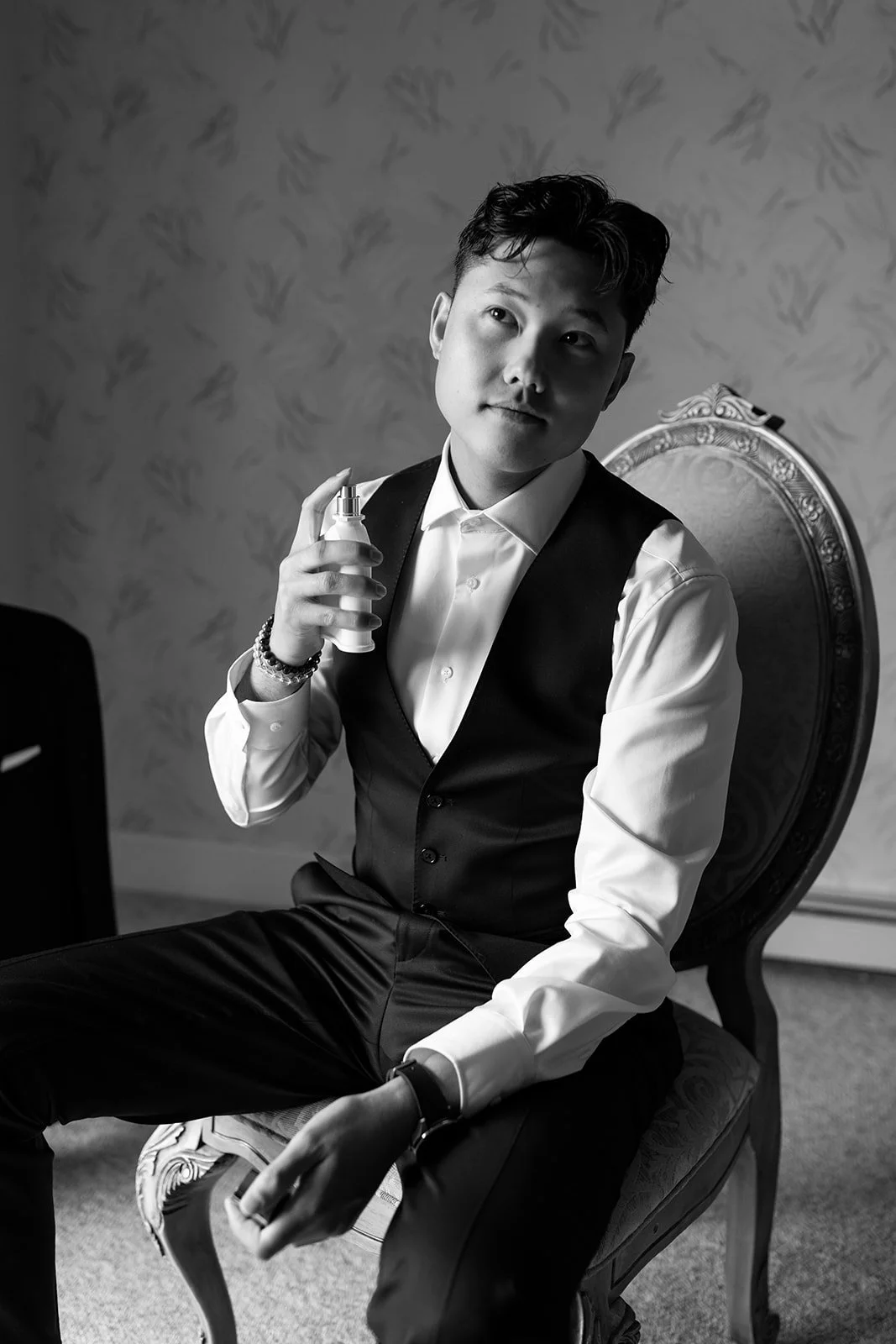 A young man dressed in formal attire sitting on an ornate chair, holding a spray bottle, looking thoughtfully to the side in a black and white portrait.