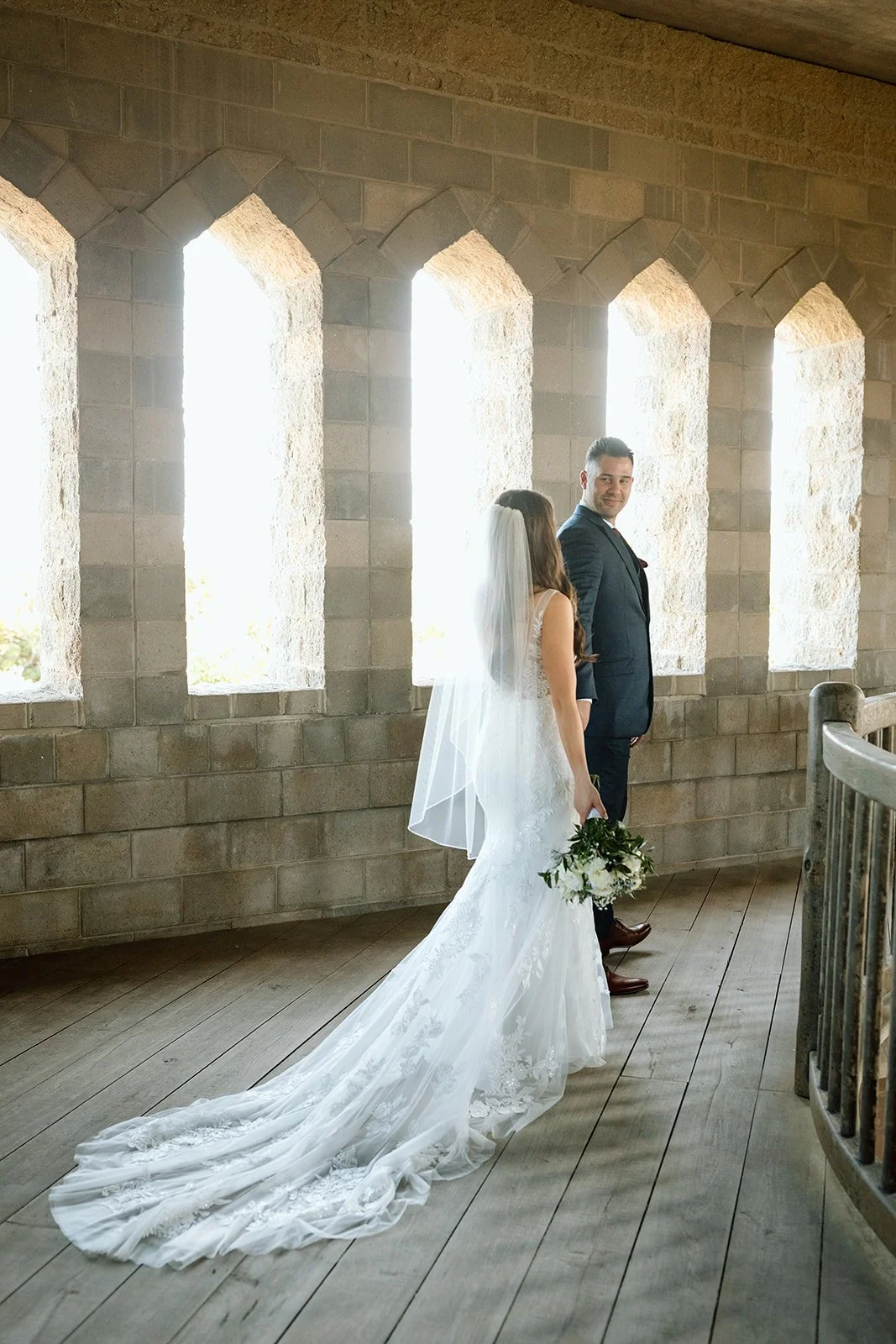 A bride and groom standing close together inside a building with stone walls and tall arched windows, with the bride holding a bouquet of flowers and wearing a white wedding dress with a long train and veil, and the groom in a dark suit, smiling at e