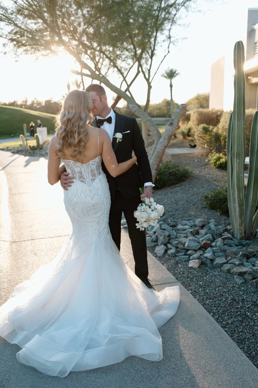 Bride and groom sharing a kiss outdoors during sunset, bride in a white wedding gown and groom in a tuxedo, with desert landscaping and cacti around them.