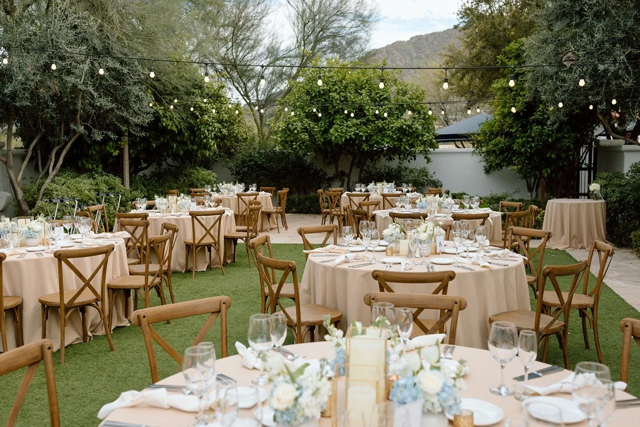Outdoor wedding reception setup with round tables covered in beige tablecloths, decorated with white and blue floral centerpieces, set with glassware and silverware, surrounded by wooden chairs, under string lights, with trees and mountains in the ba