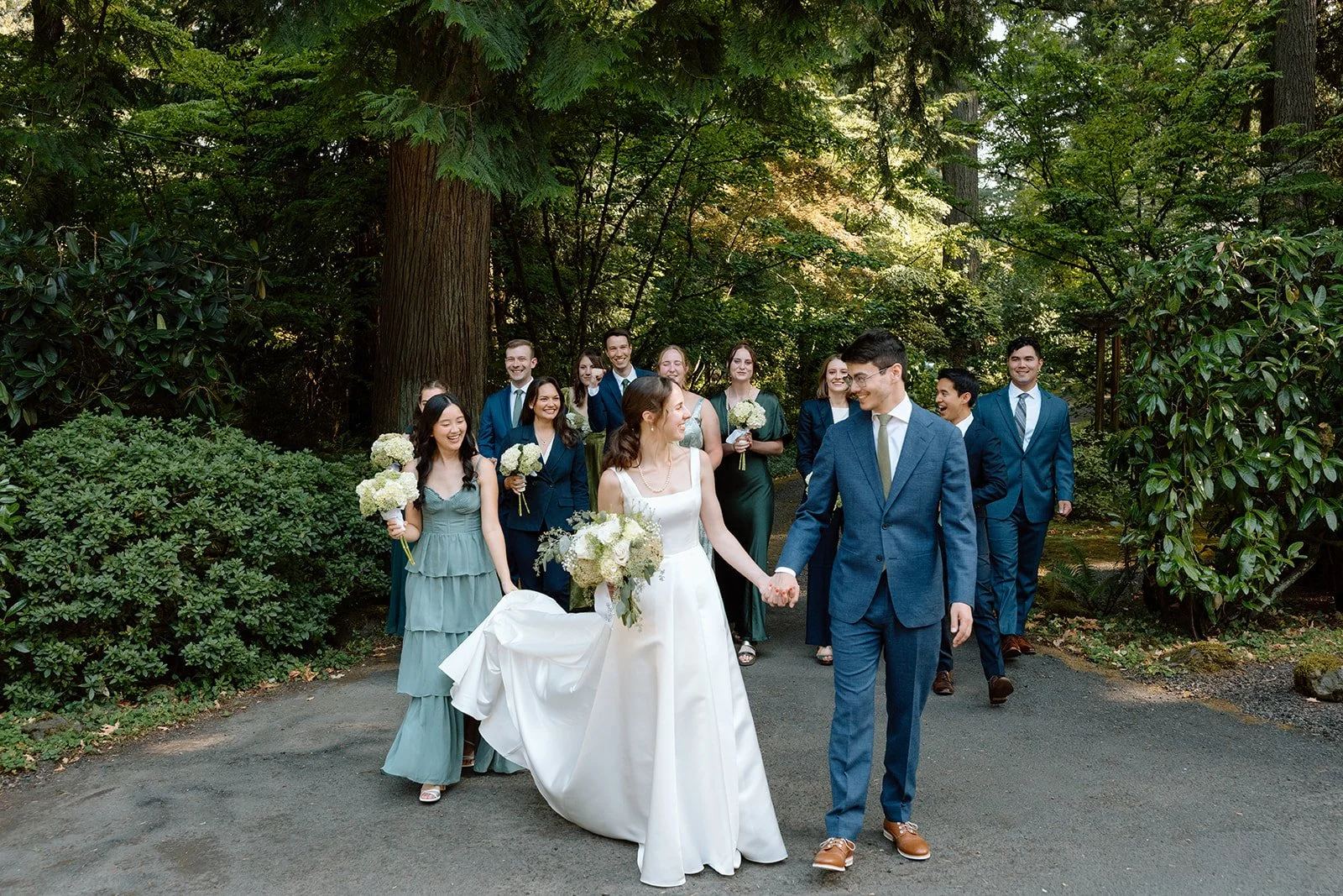 A wedding party walking outdoors on a paved path surrounded by trees and greenery. The bride and groom are in the forefront, holding hands and smiling at each other. The bride wears a white wedding dress and holds a bouquet, while the groom wears a b