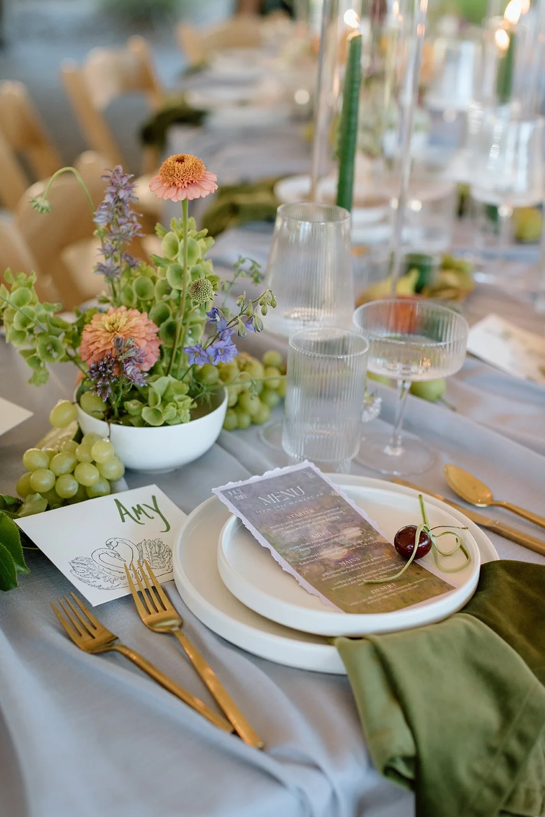 Elegant table setting with a floral centerpiece, green grapes, gold utensils, a menu card, a Cherry, and a personalized place card reading 'Amy.'