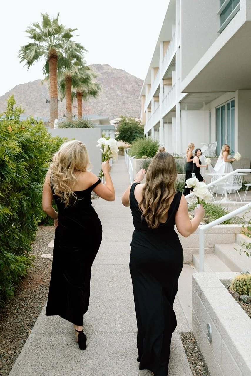 Women in black dresses walking on a sidewalk, carrying white flowers, at a wedding ceremony outdoors with palm trees and mountains in the background.