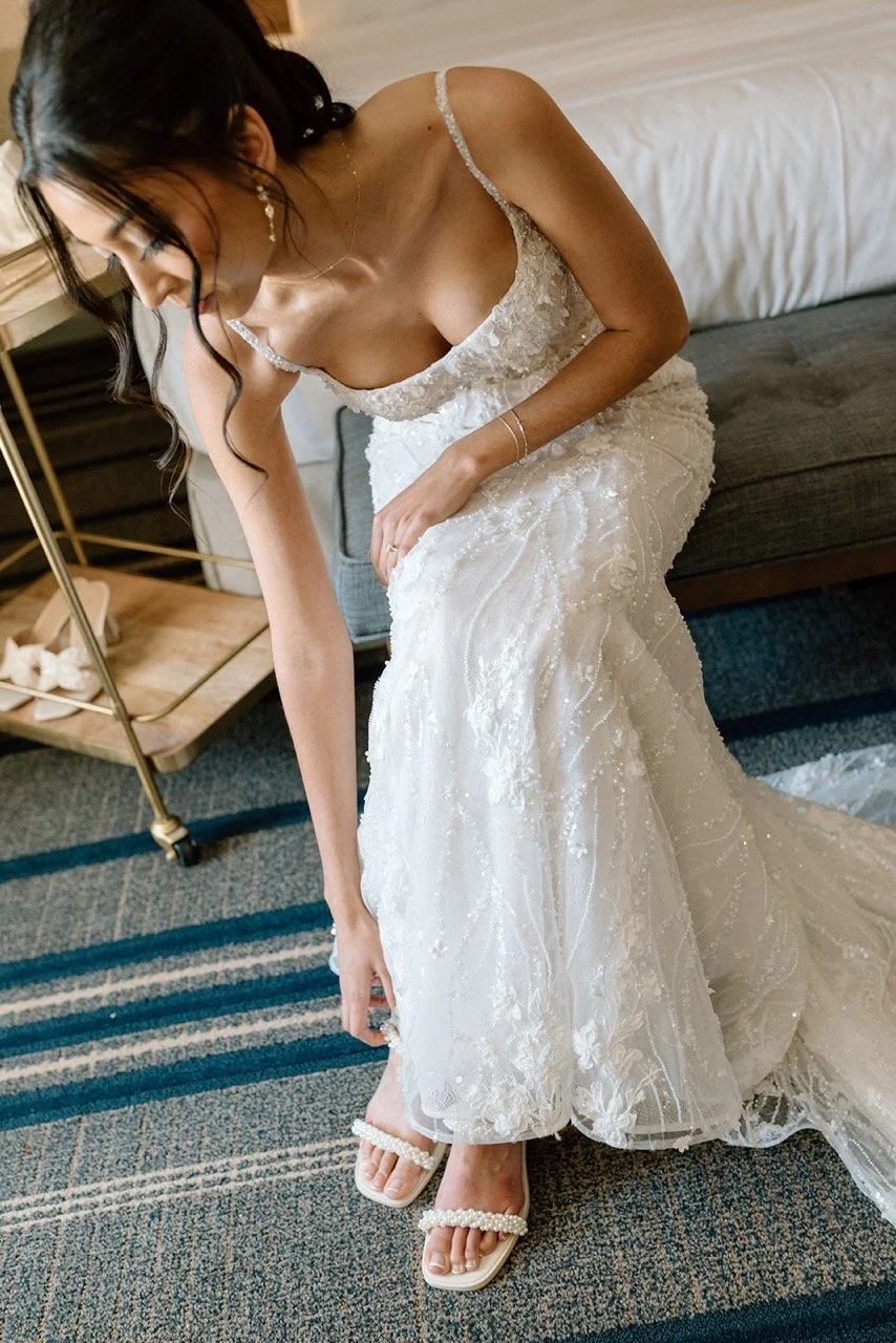 Brunette bride in a white lace wedding gown adjusting her shoe in a hotel room.