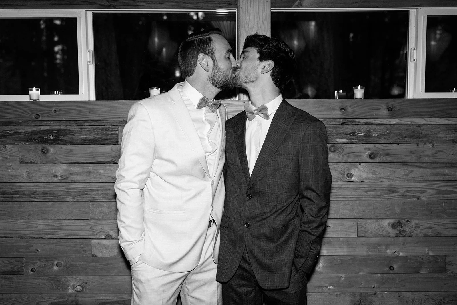 Two men in tuxedos kissing in front of a wooden wall with candles in the background.