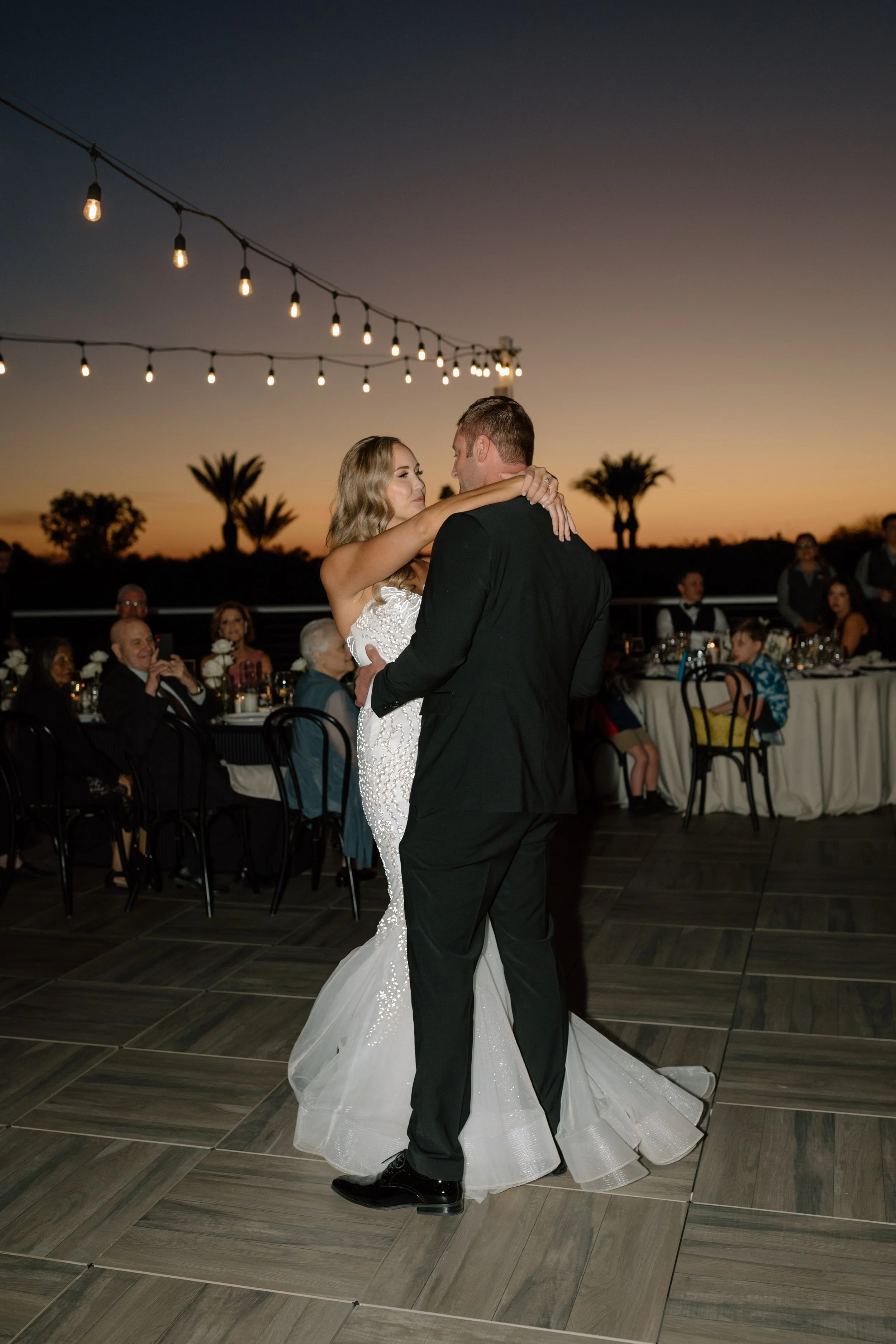 A bride and groom sharing their first dance during sunset at their wedding reception, with guests sitting at round tables in the background and string lights overhead.