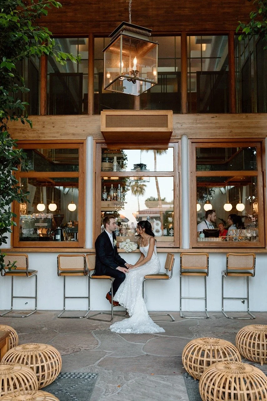 A bride and groom sitting closely and holding hands at a bar area with stairs above, surrounded by wicker stools, trees, and windows reflecting a sunset sky.