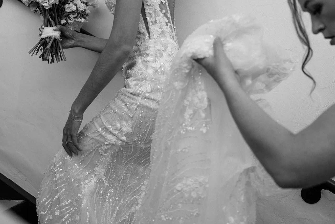 A woman in an wedding dress holding a bouquet while another woman adjusts the dress.