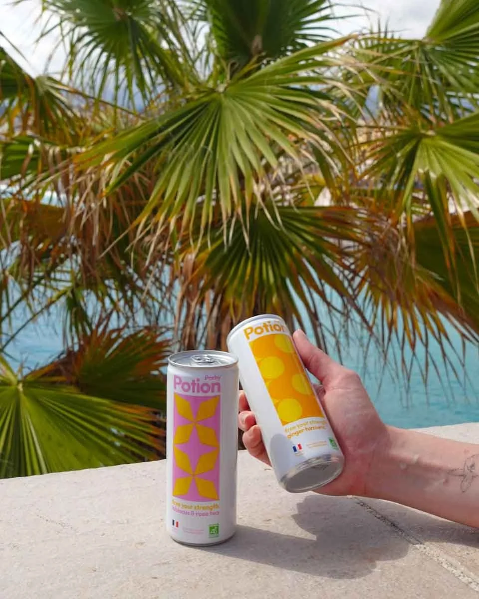 Deux canettes de boisson énergisante intitulées 'Potion' en main d'une personne contre un fond de palmiers