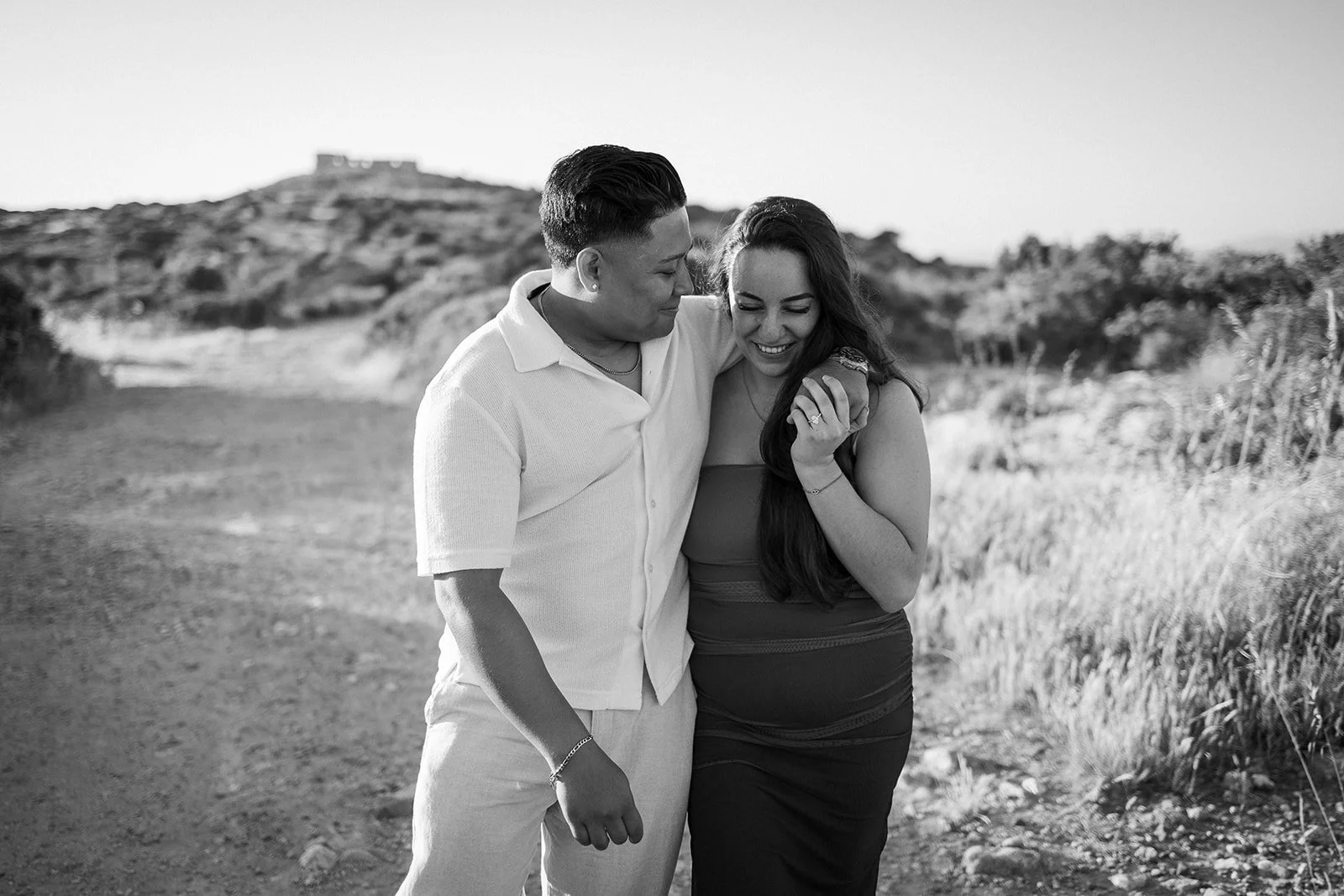 A black and white photo of a couple smiling and embracing outdoors, with a hill and a building in the background.