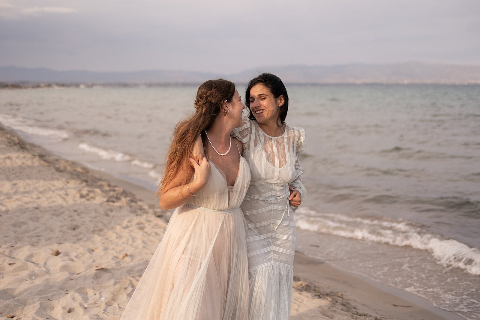 Two women in white dresses smiling and embracing each other on the beach near the water.