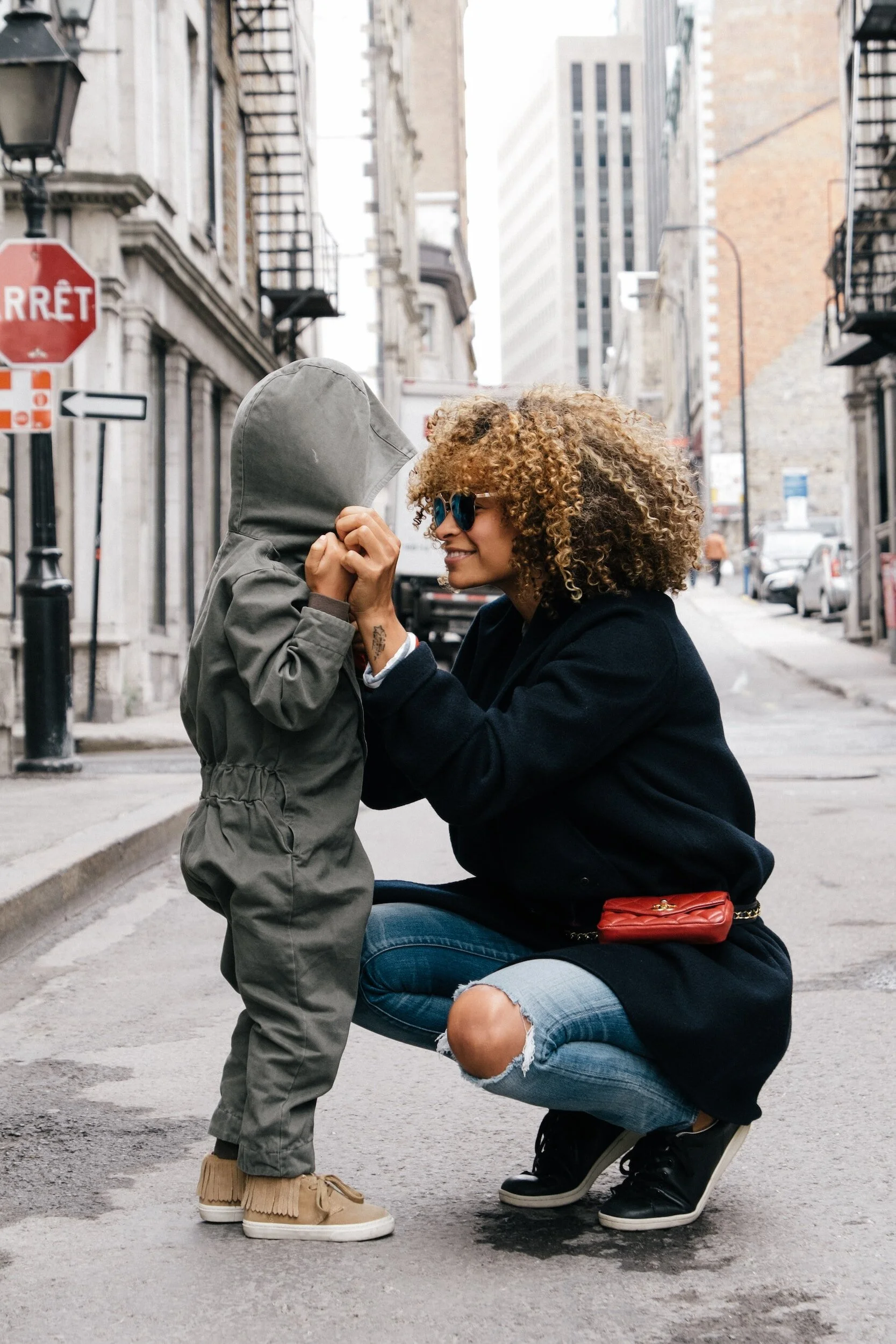 A woman crouching and smiling at a young child who is standing on a city street, with buildings and cars in the background.
