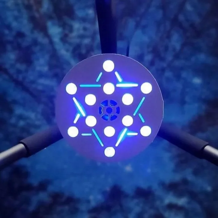 Close-up of a circular device with blue LED lights connecting white dots, forming a geometric pattern, attached to a thopter with propellers visible on each side, set against a cloudy blue sky.