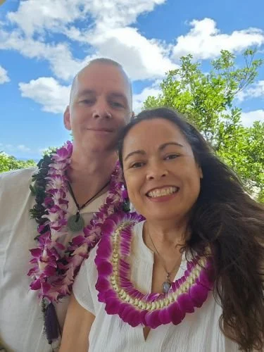 A man and a woman smiling outdoors with blue sky and green trees in the background, both wearing leis.