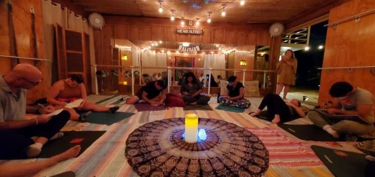 People sitting on yoga mats in a wooden room with string lights, candles, and a 'We Are Blessed' sign, participating in a yoga or meditation session at night.