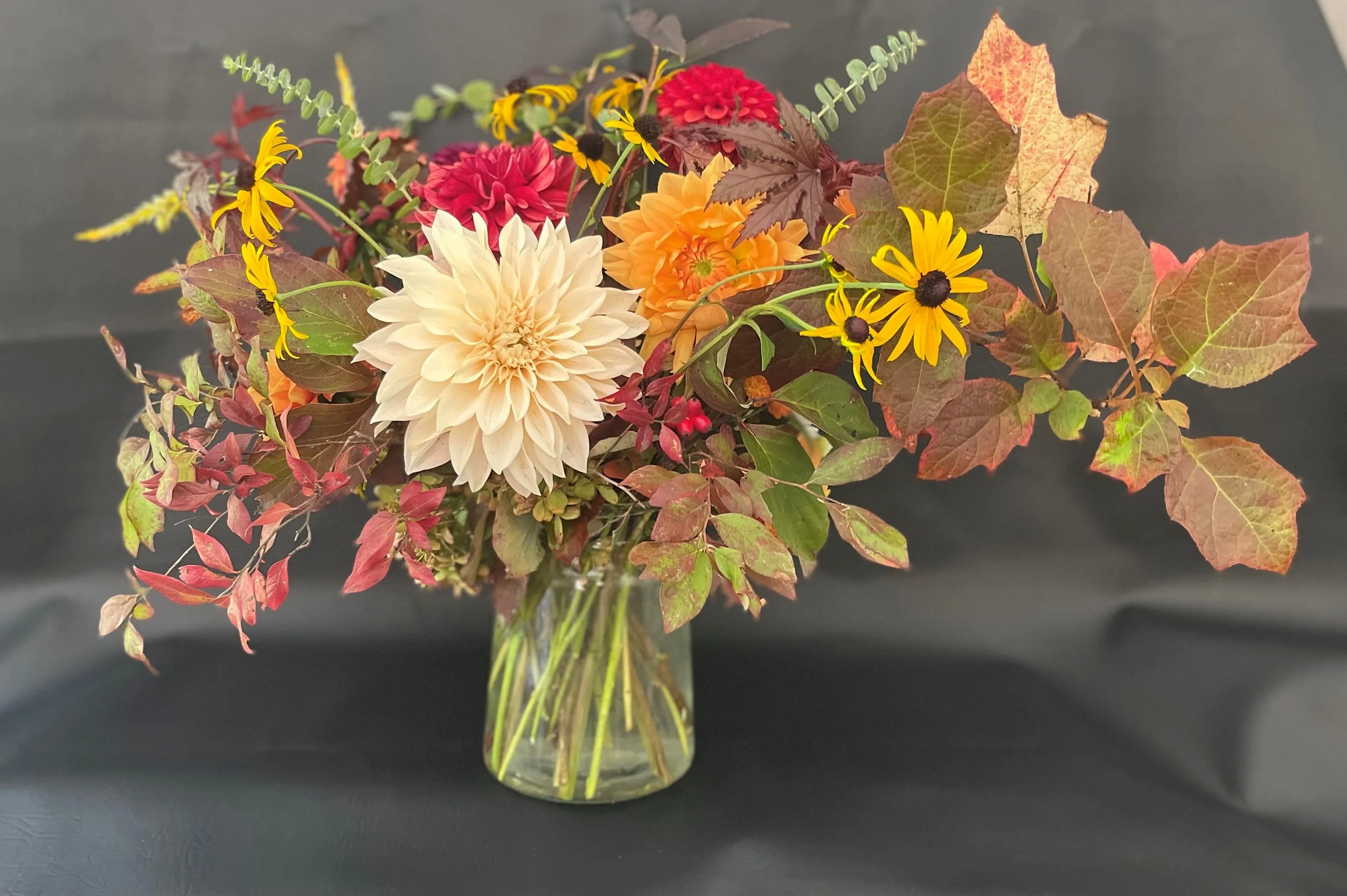 Autumn floral arrangement in a clear glass vase with various colorful flowers and fall leaves, including white, pink, orange, and yellow blooms against a dark background.