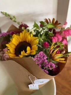 Bouquet of colorful flowers including a sunflower, pink blooms, and purple flowers wrapped in brown paper.