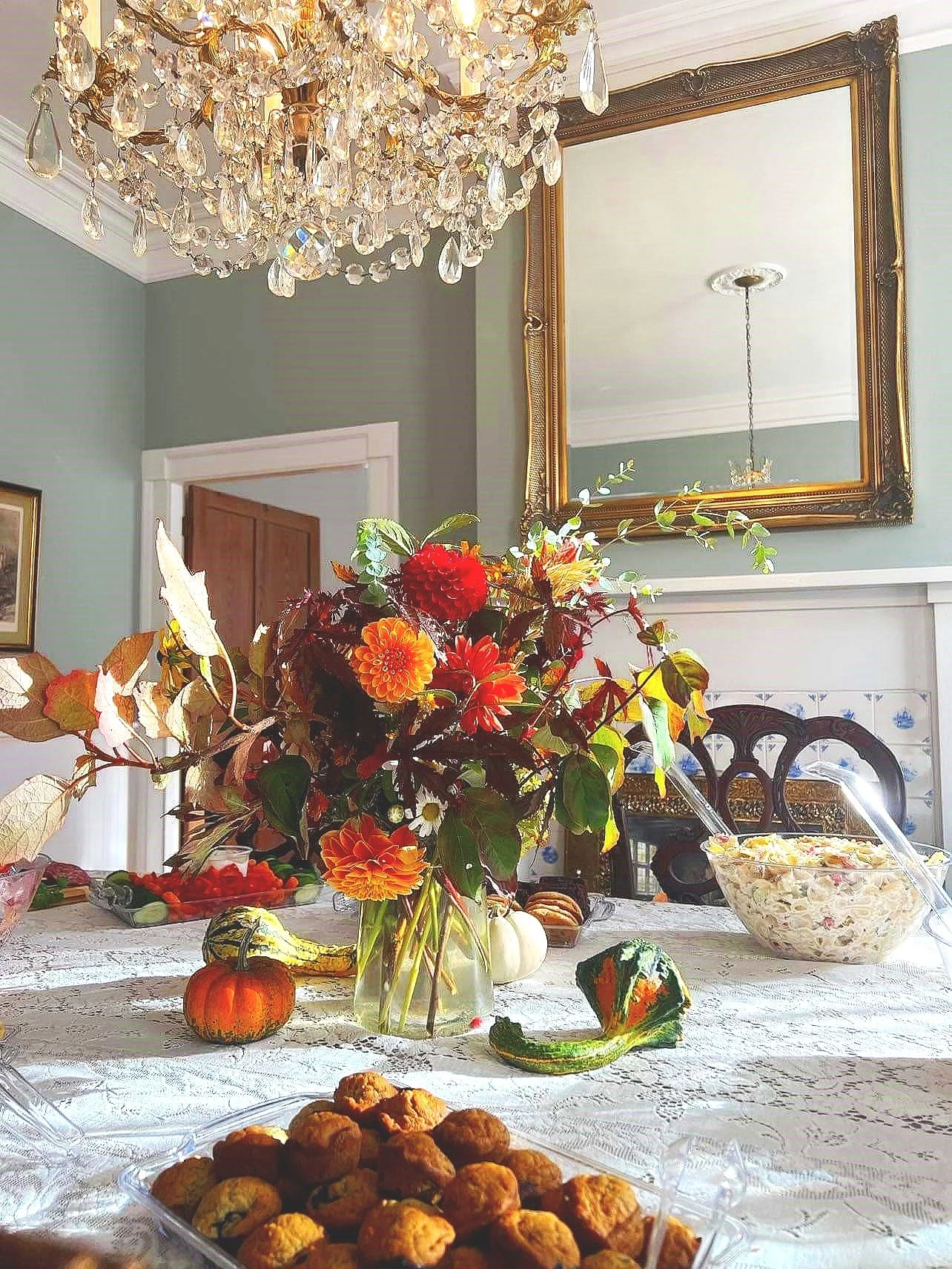 A dining room table with a floral arrangement, pumpkins, squash, and a tray of baked goods, with a chandelier overhead and a large mirror on the wall.
