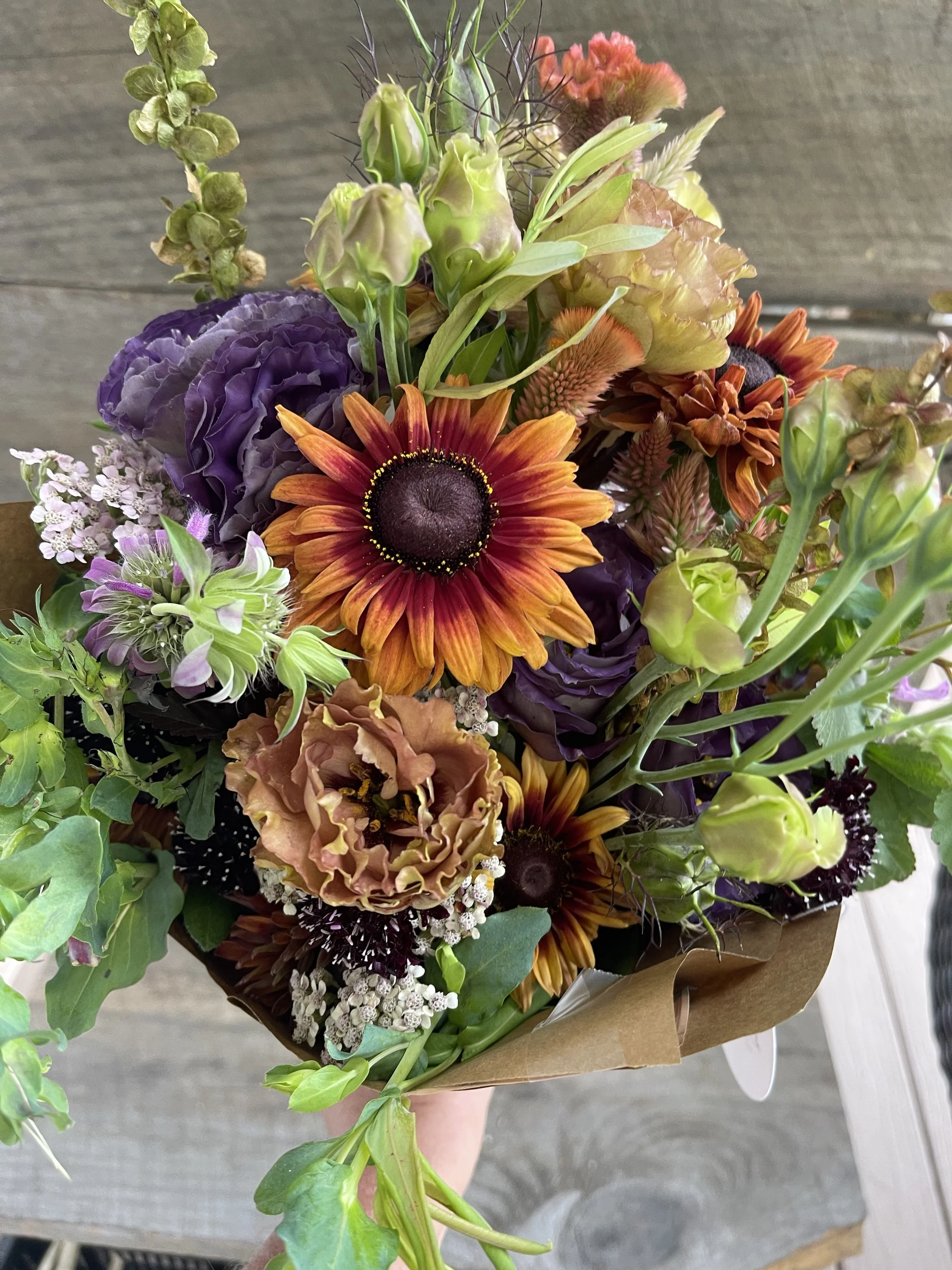 Colorful bouquet of various flowers including orange and red sunflowers, purple carnations, green buds, and small white and purple filler flowers, wrapped in brown paper.