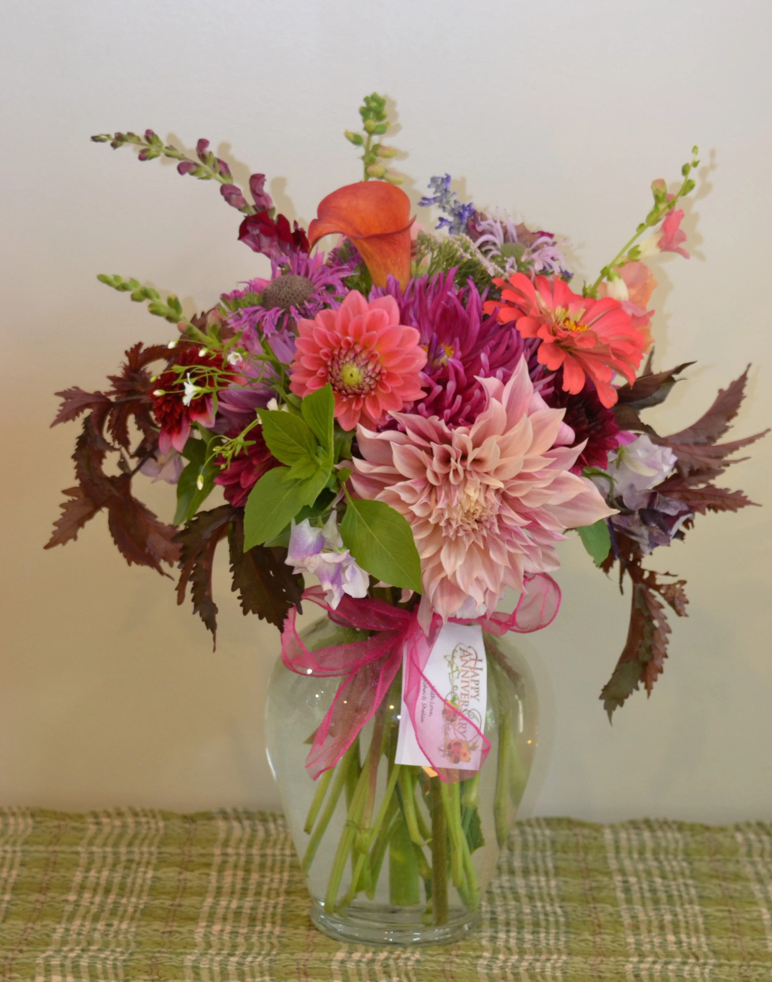 A colorful bouquet of various flowers in a clear glass vase on a woven tablecloth.