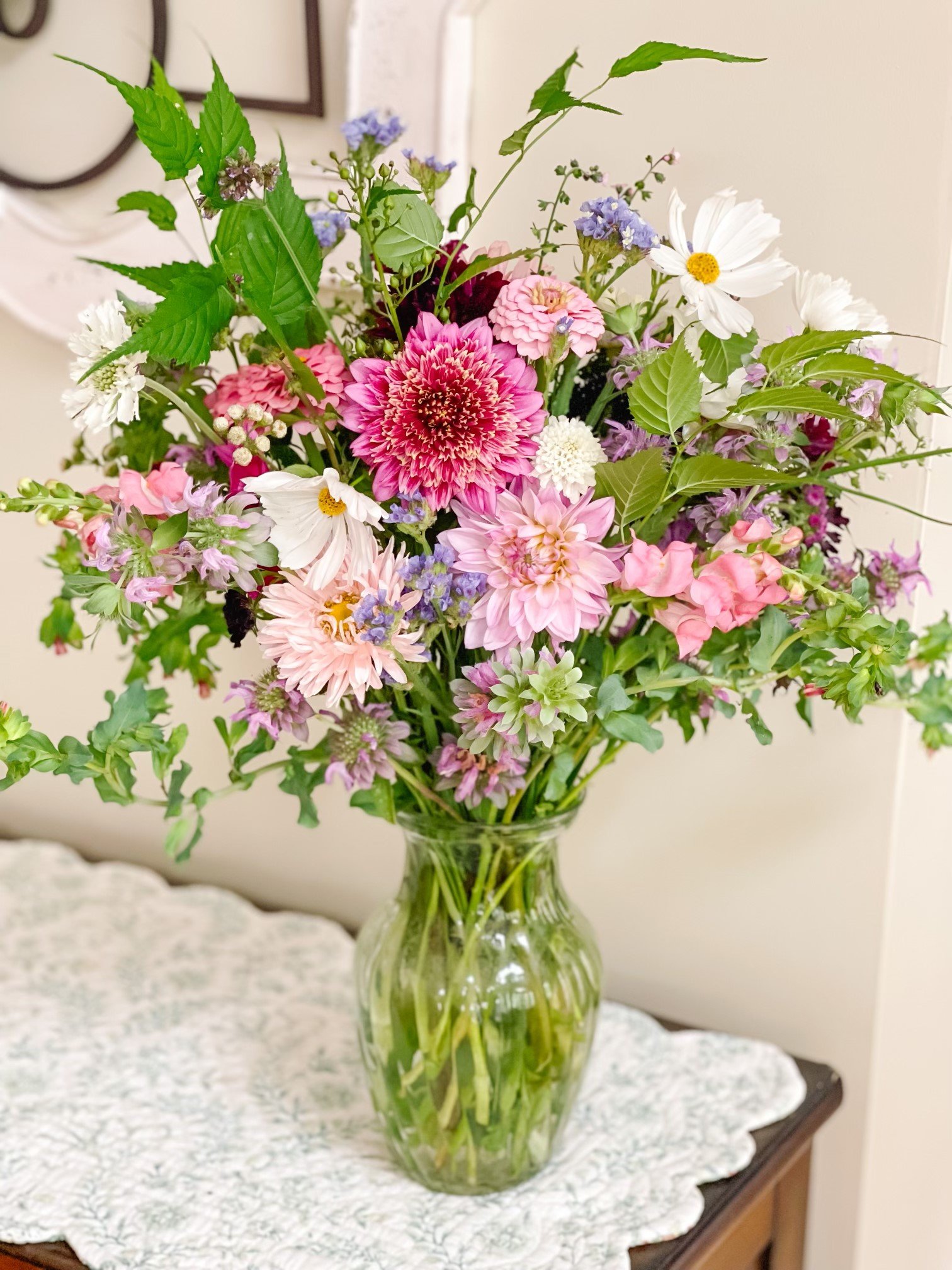 A glass vase filled with various colorful flowers including pink, purple, white, and green blossoms on a table with a crocheted white doily.