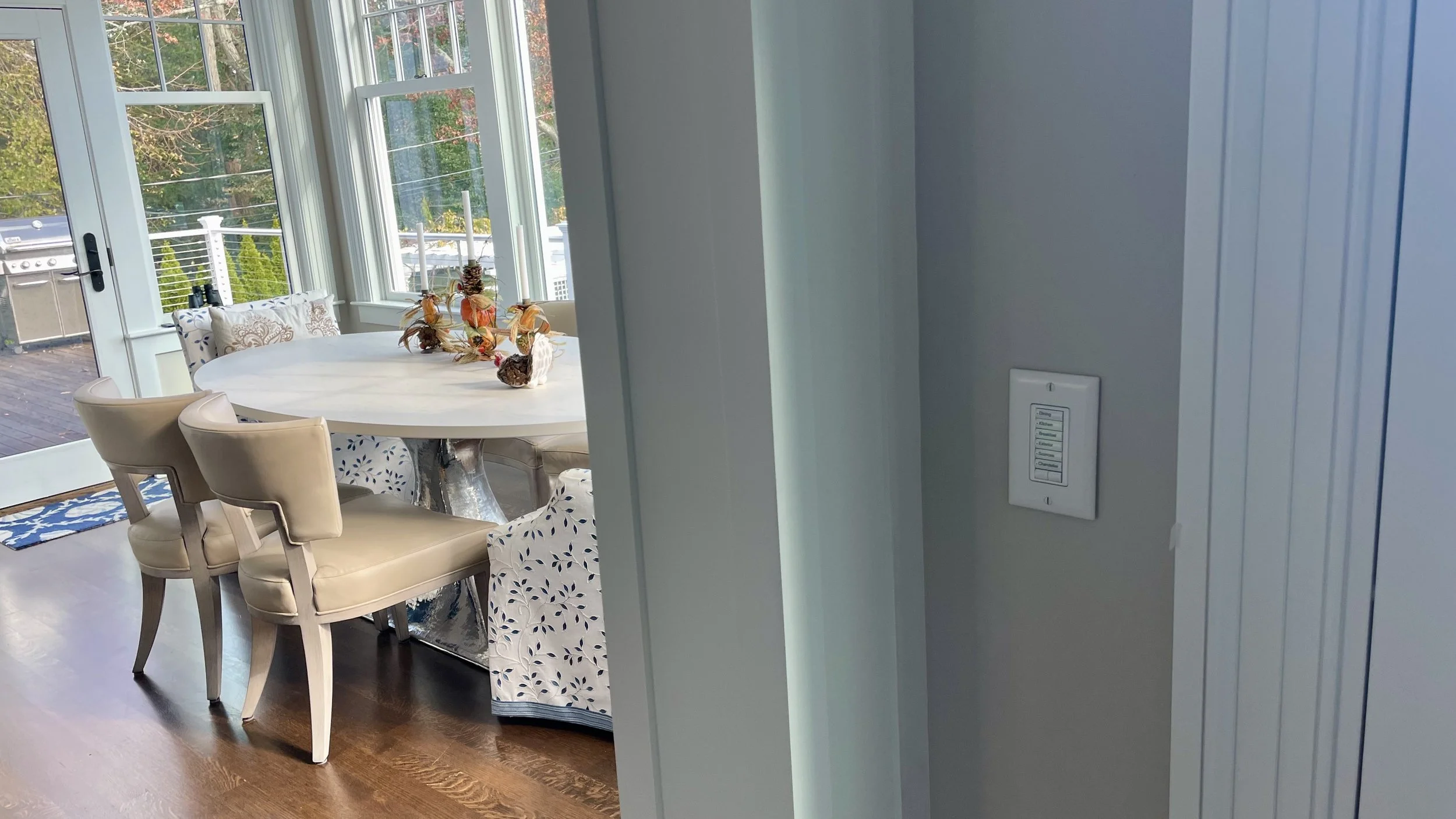 A dining room with a round table decorated with autumn-themed centerpiece and surrounded by upholstered chairs. Large windows provide a view of the outdoor deck and greenery.