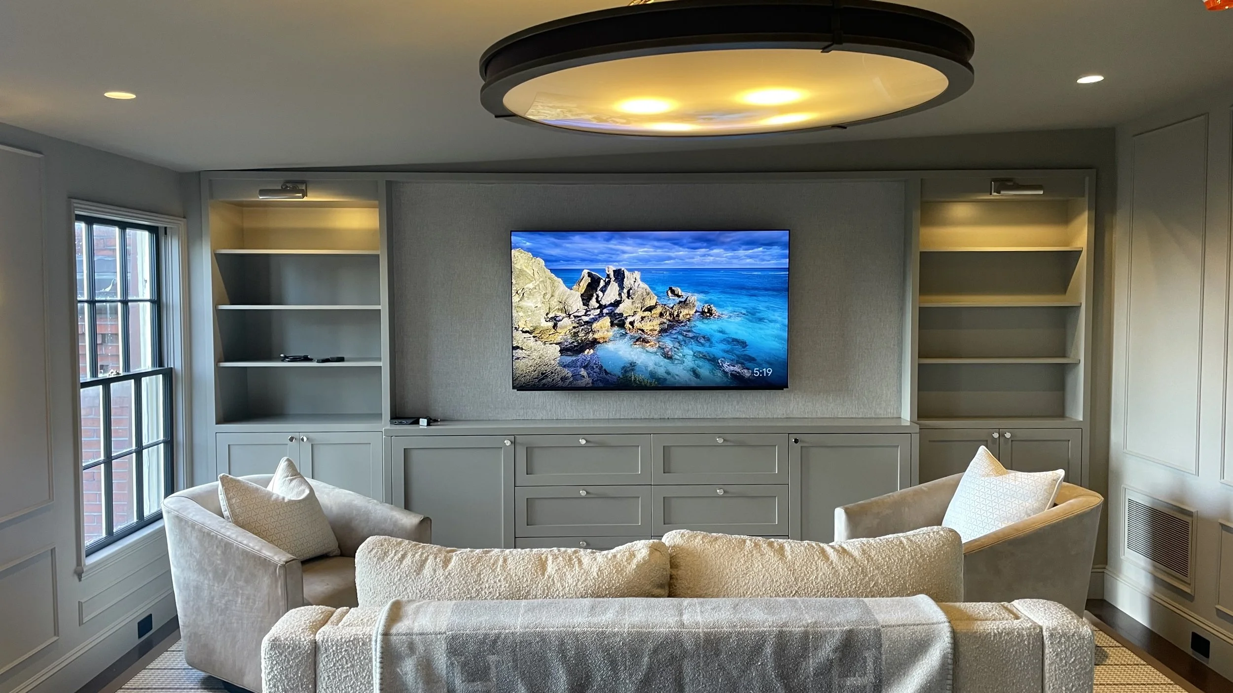 Living room with a wall-mounted television displaying a rocky coastline scene, surrounded by built-in shelves and cabinets, with two armchairs and a sofa.