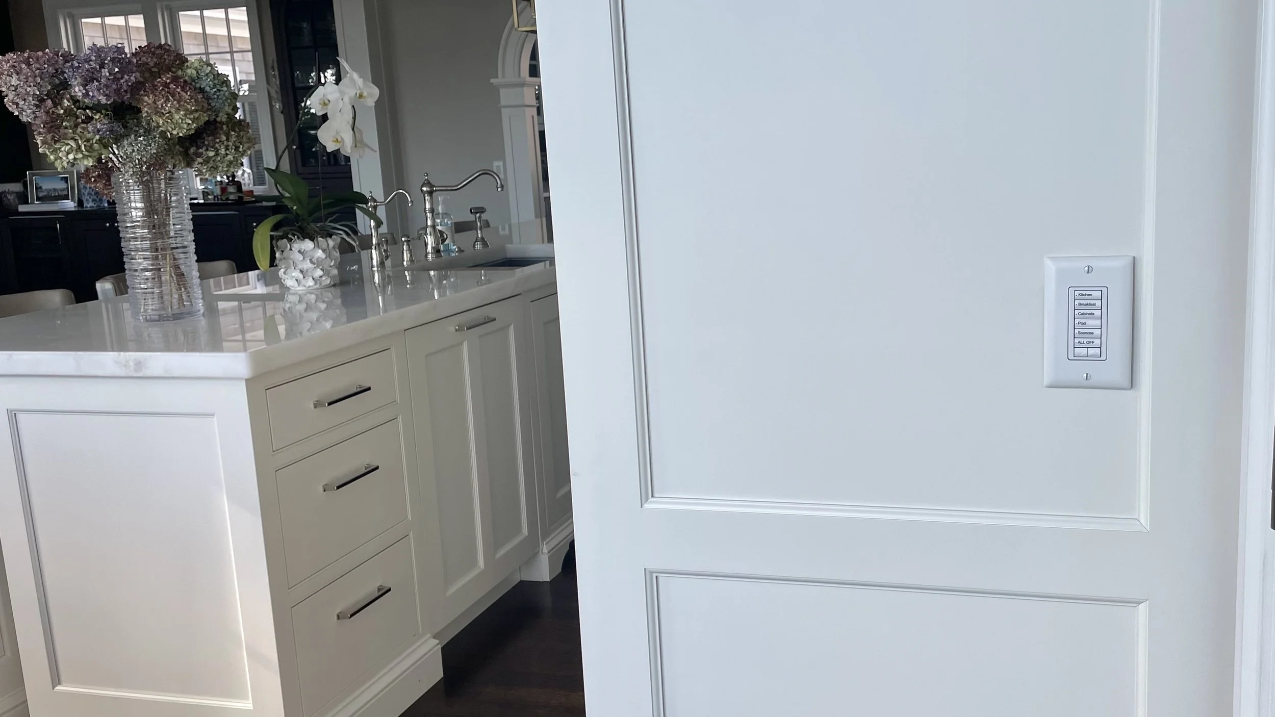 White kitchen island with a sink, adorned with large flower arrangements in vases. A wall with a switch and the kitchen is visible in the background.