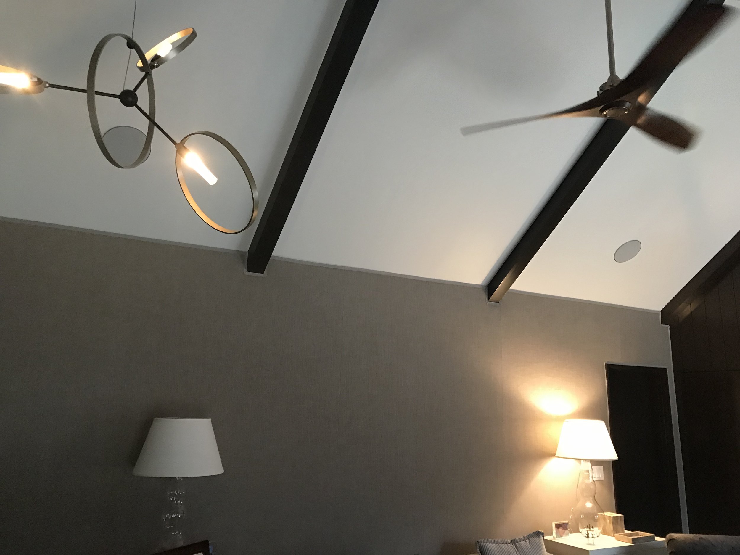 A ceiling with a modern chandelier, a ceiling fan, a wall with a lamp, and a side table with a lamp and some decorative items.