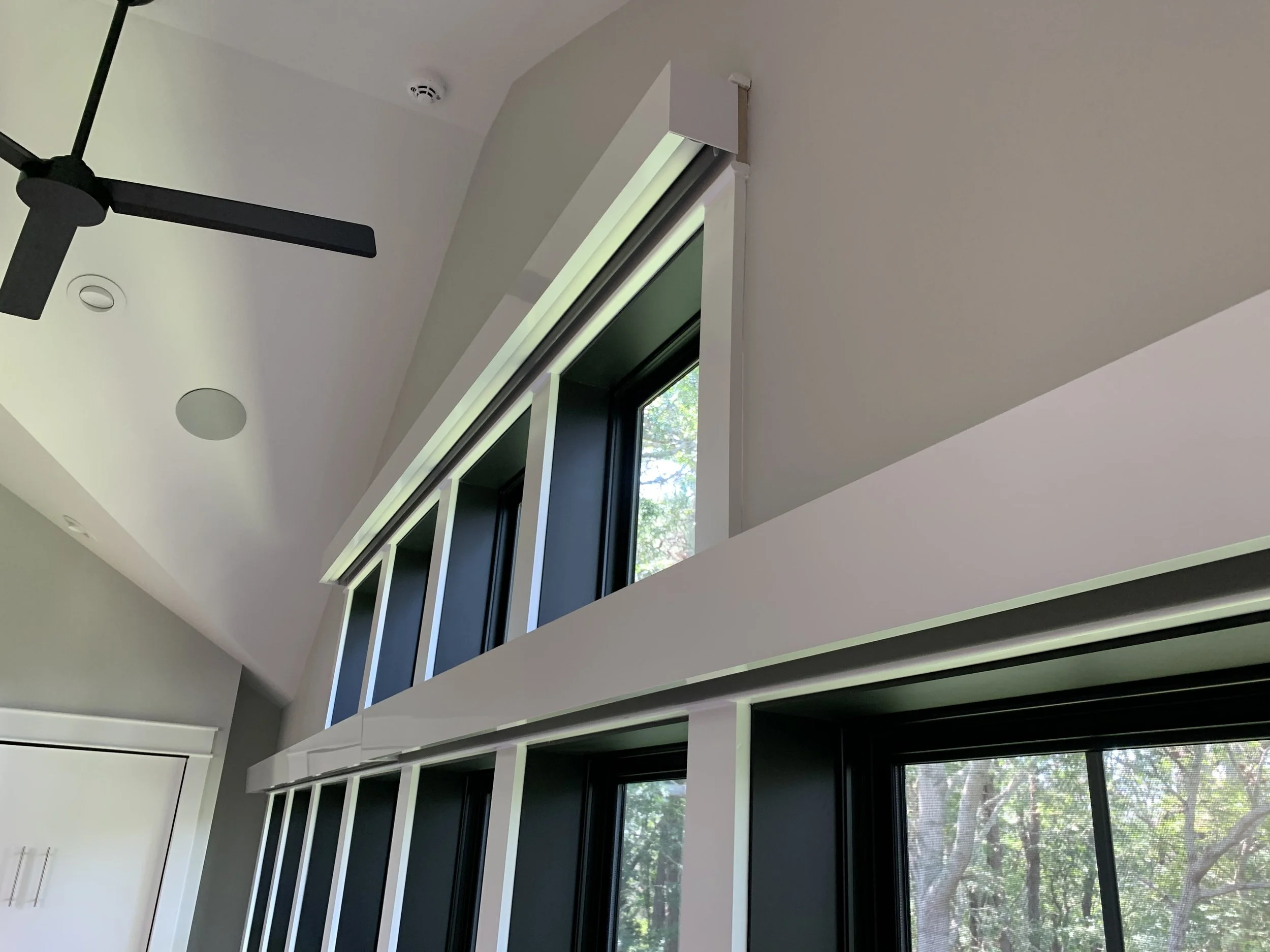 Interior view of a modern room with tall windows, black window frames, a white ceiling with a black ceiling fan, and Lutron Triathalon shades.