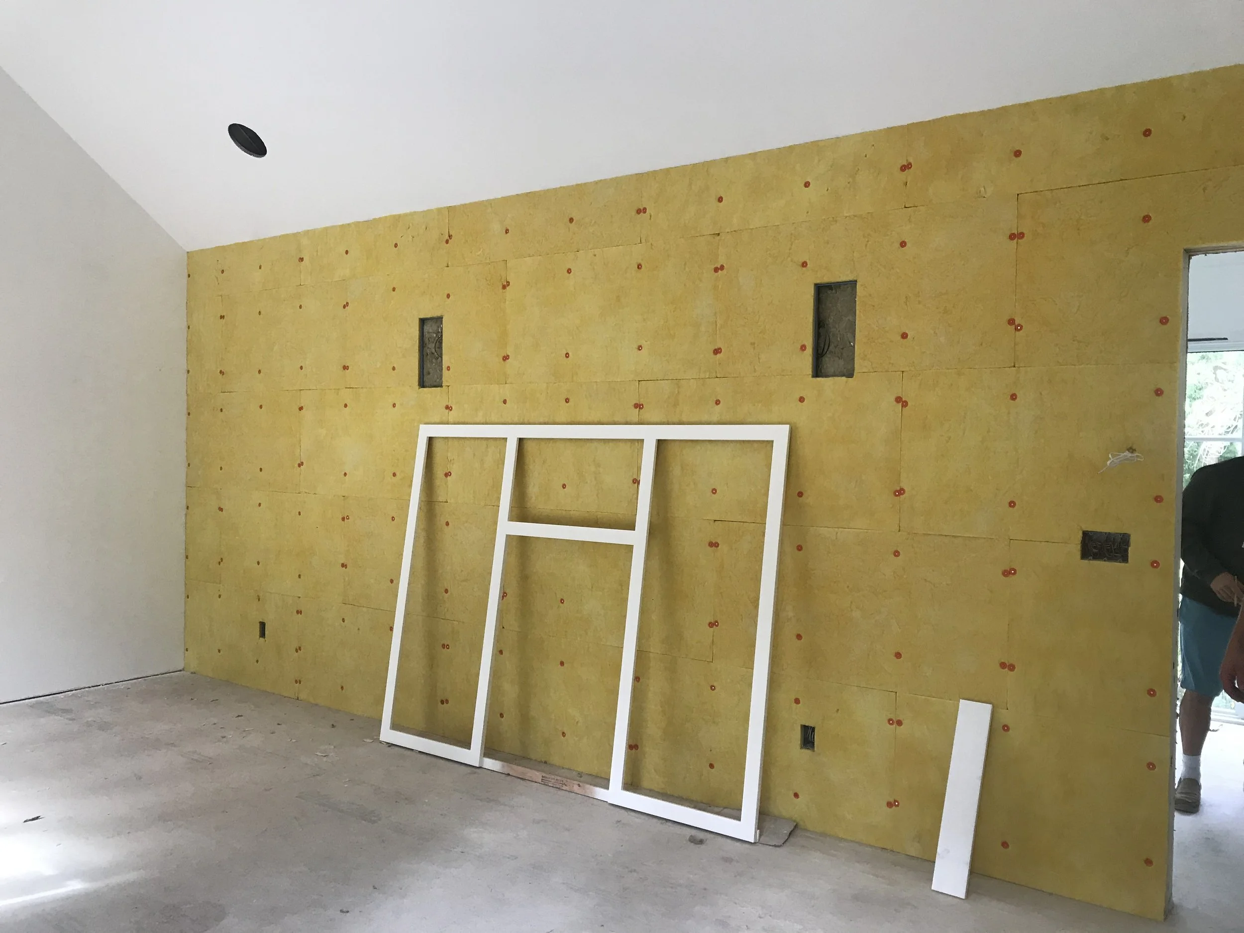 Interior of a room under construction with a yellow wall made of insulation or drywall panels, visible cutouts and electrical outlets, a white frame leaning against the wall, and a person partially visible in the doorway.