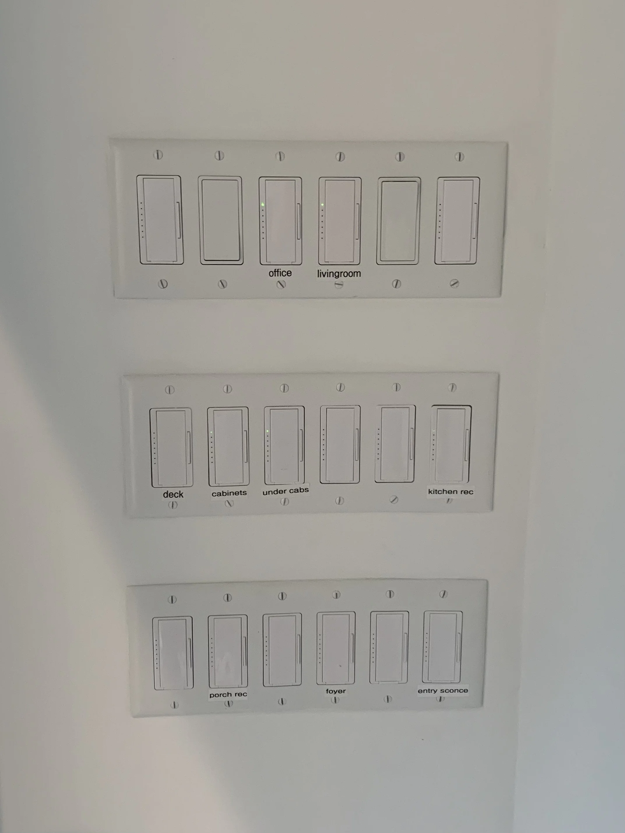 A wall with three rows of Lutron Pro switches labeled for different rooms and areas, including office, living room, deck, cabinets, under cabinets, kitchen, porch, foyer, and entry sconce.
