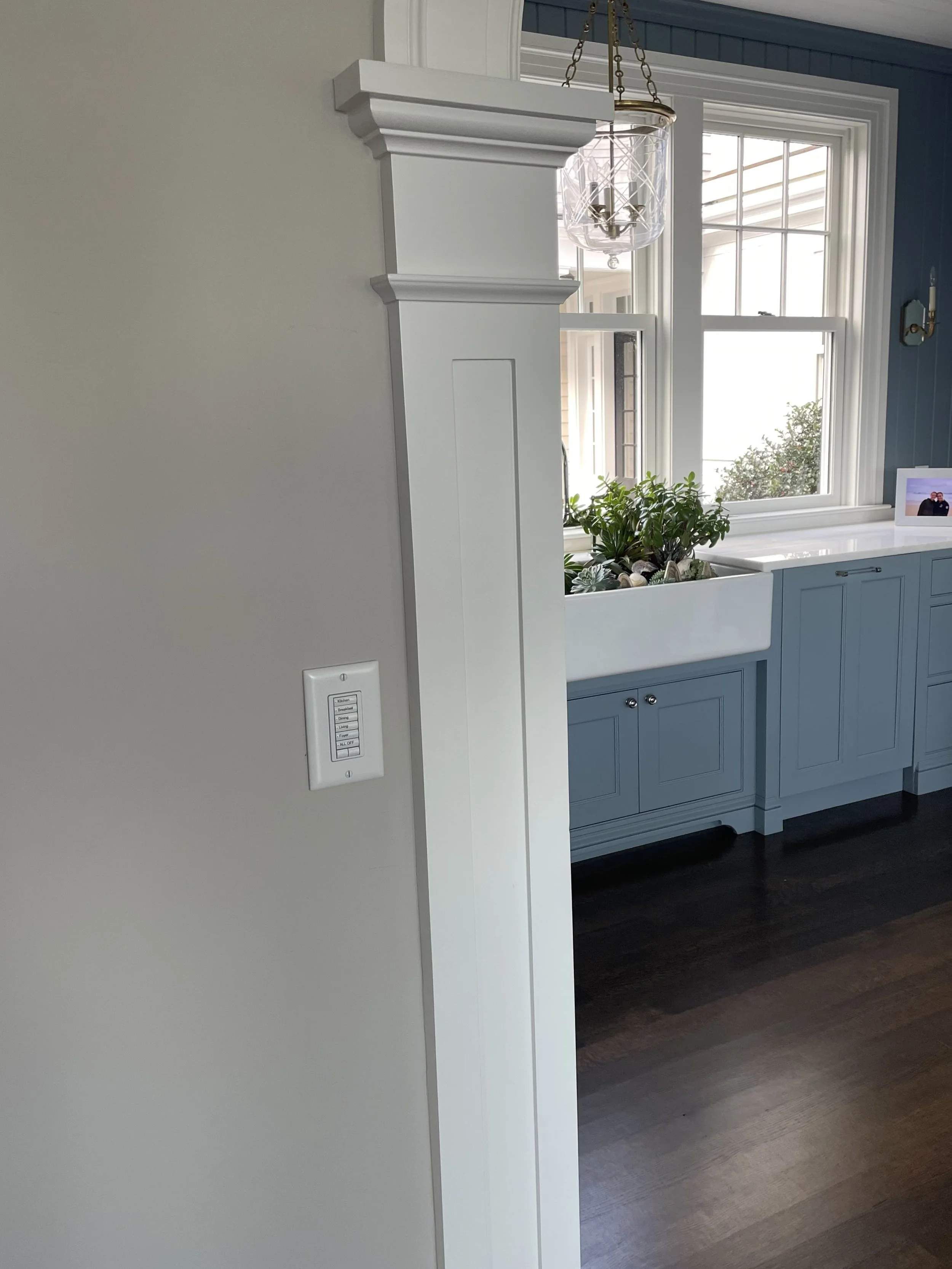 White kitchen wall with light switch, white decorative column, and a view of a window, blue cabinetry, and a planter with greenery and rocks inside.
