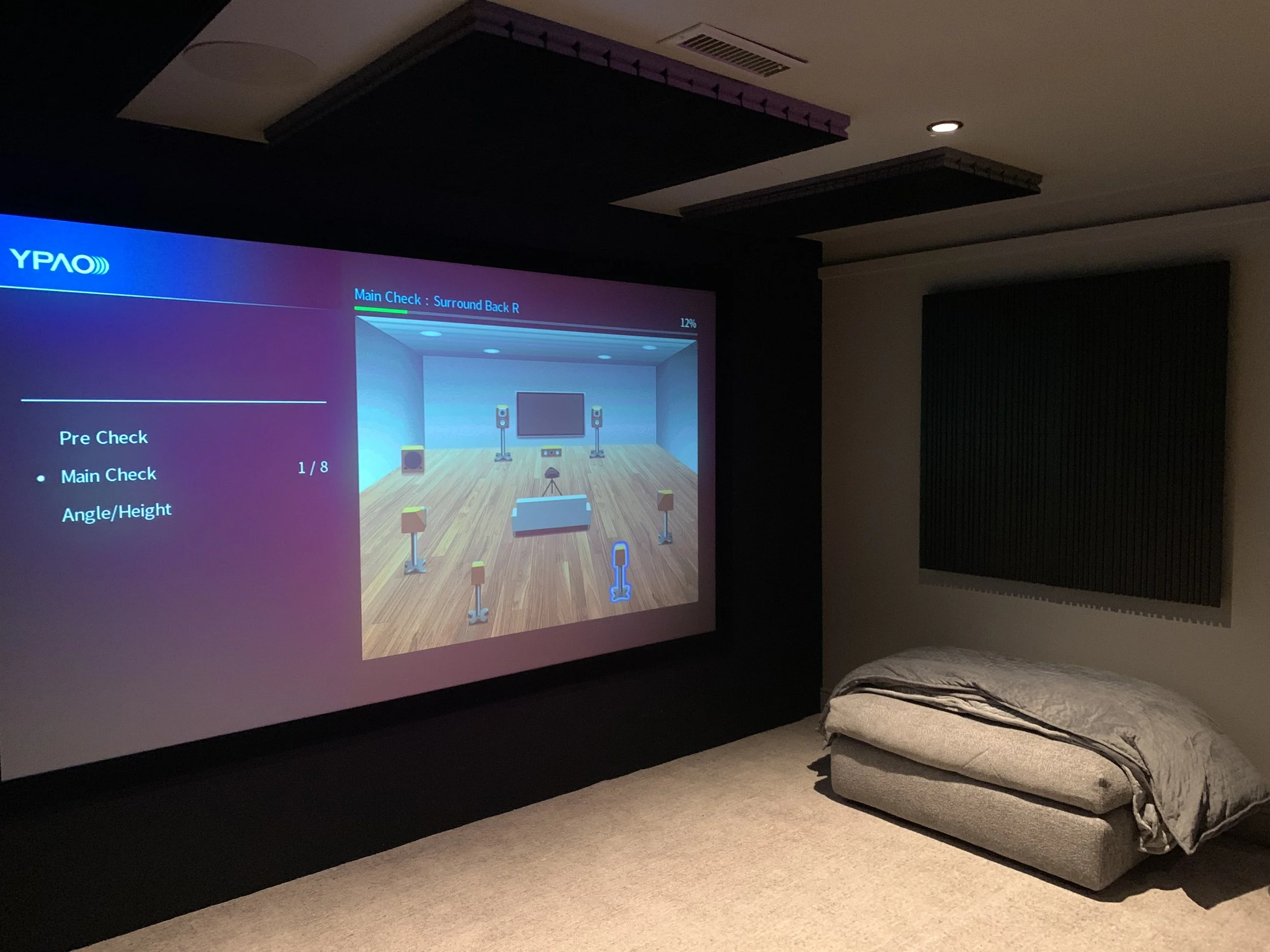 A home theater room with a large projector screen on the left showing speaker setup instructions, a folded gray sofa on the right, and dark soundproofing panels on the wall. The room has ceiling lights and a cozy carpeted floor.