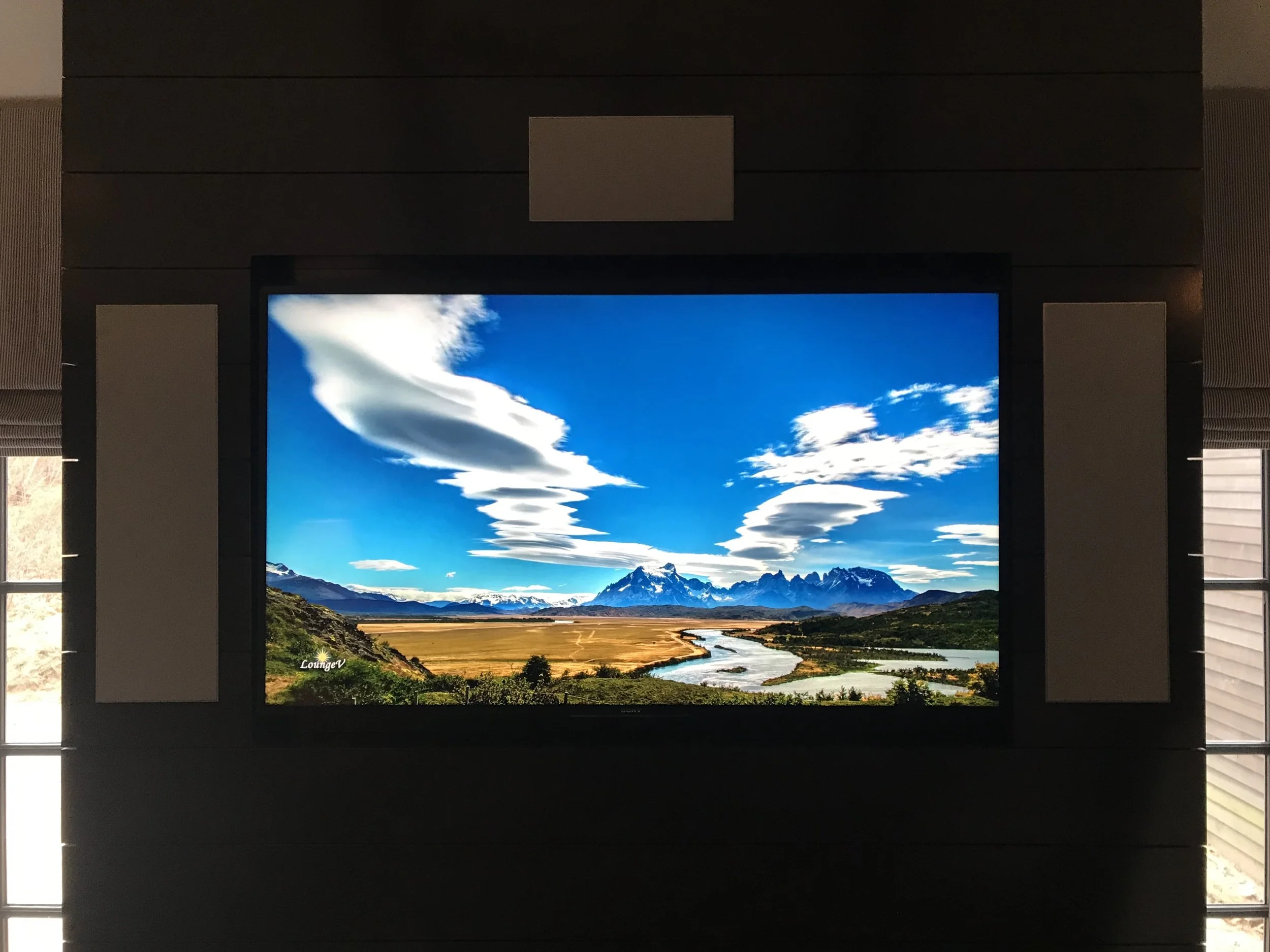 A landscape of mountains, river, and a blue sky with unique white cloud formations displayed on a large screen in a room.