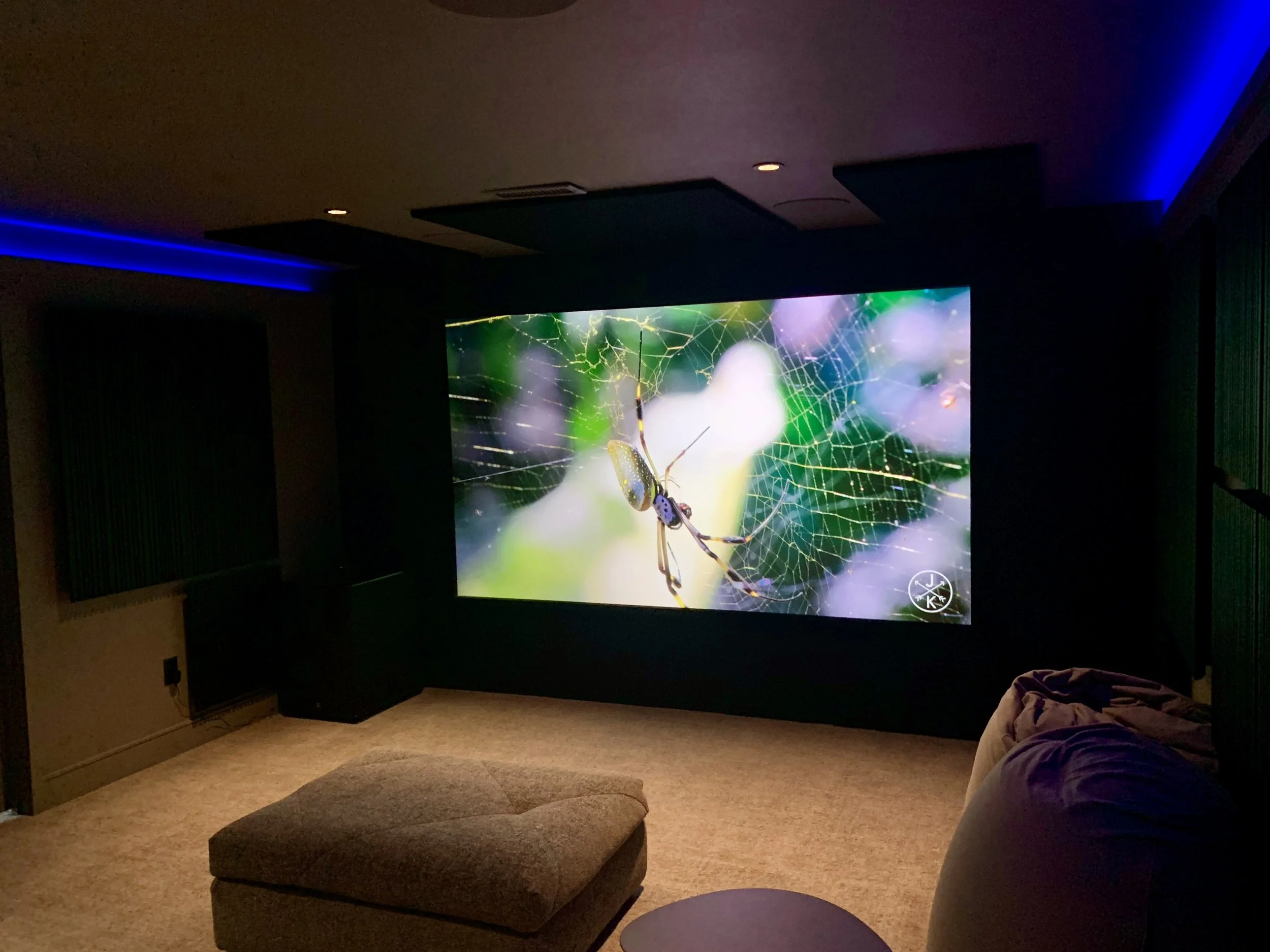 A home theater room with a large screen displaying a close-up of a spider on its web. There is a small ottoman, a reclining chair, and some acoustic panels on the walls, with ambient blue lighting.