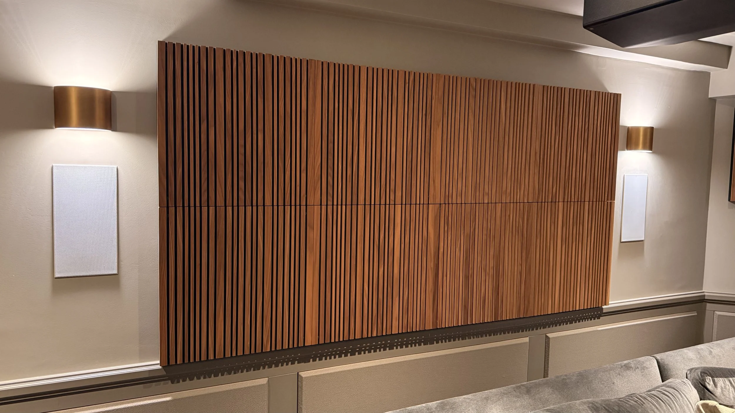 Interior of a room with beige walls and a wooden slat wall feature, two wall-mounted lights, and a grey upholstered piece of furniture at the bottom right corner.