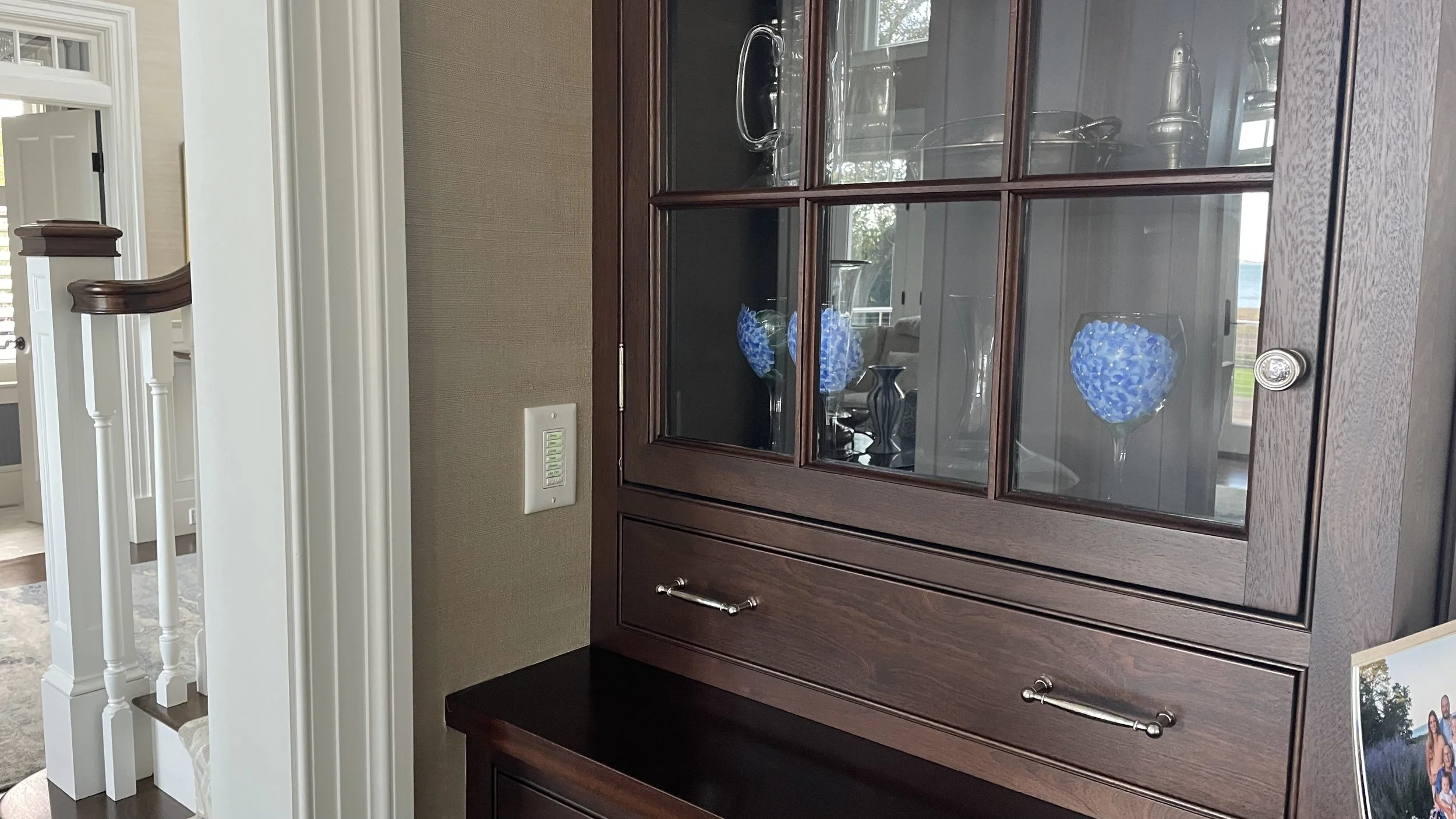 A wooden display cabinet with glass doors showcasing blue floral arrangements inside. The cabinet has a lower drawer with a metal handle, and is positioned near a beige wall with a light switch. Part of a staircase and a door can be seen in the backg