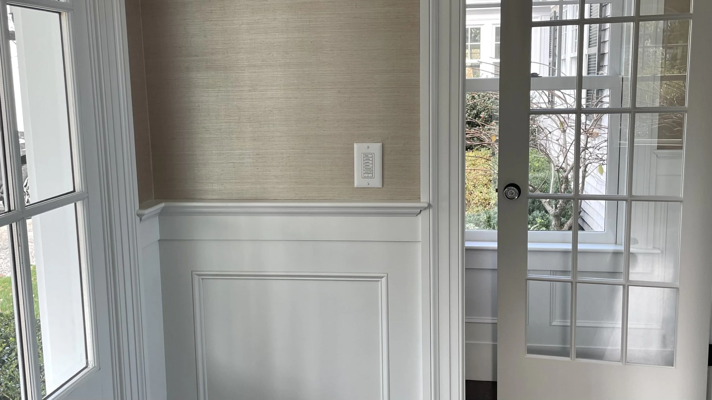 Interior view of a room with a beige textured wall, a white wainscot, a switch panel, a framed glass door, and a large window showing a garden outside.
