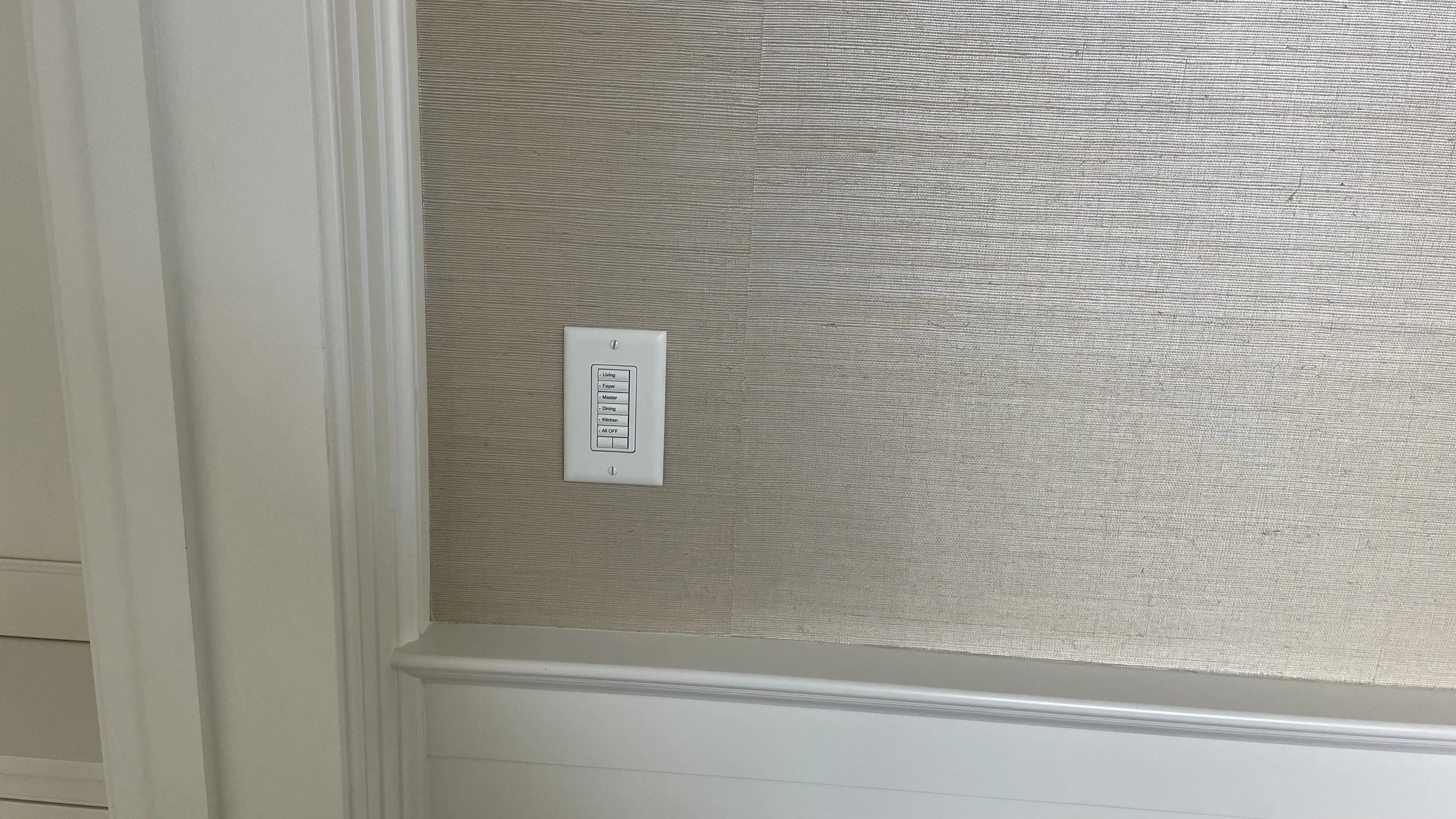 A beige wall with a light switch panel. The switch panel has several labeled switches: Living, Foyer, Master, Dining, Kitchen, and an All Off switch. There's decorative white molding at the bottom and on the left side of the wall.