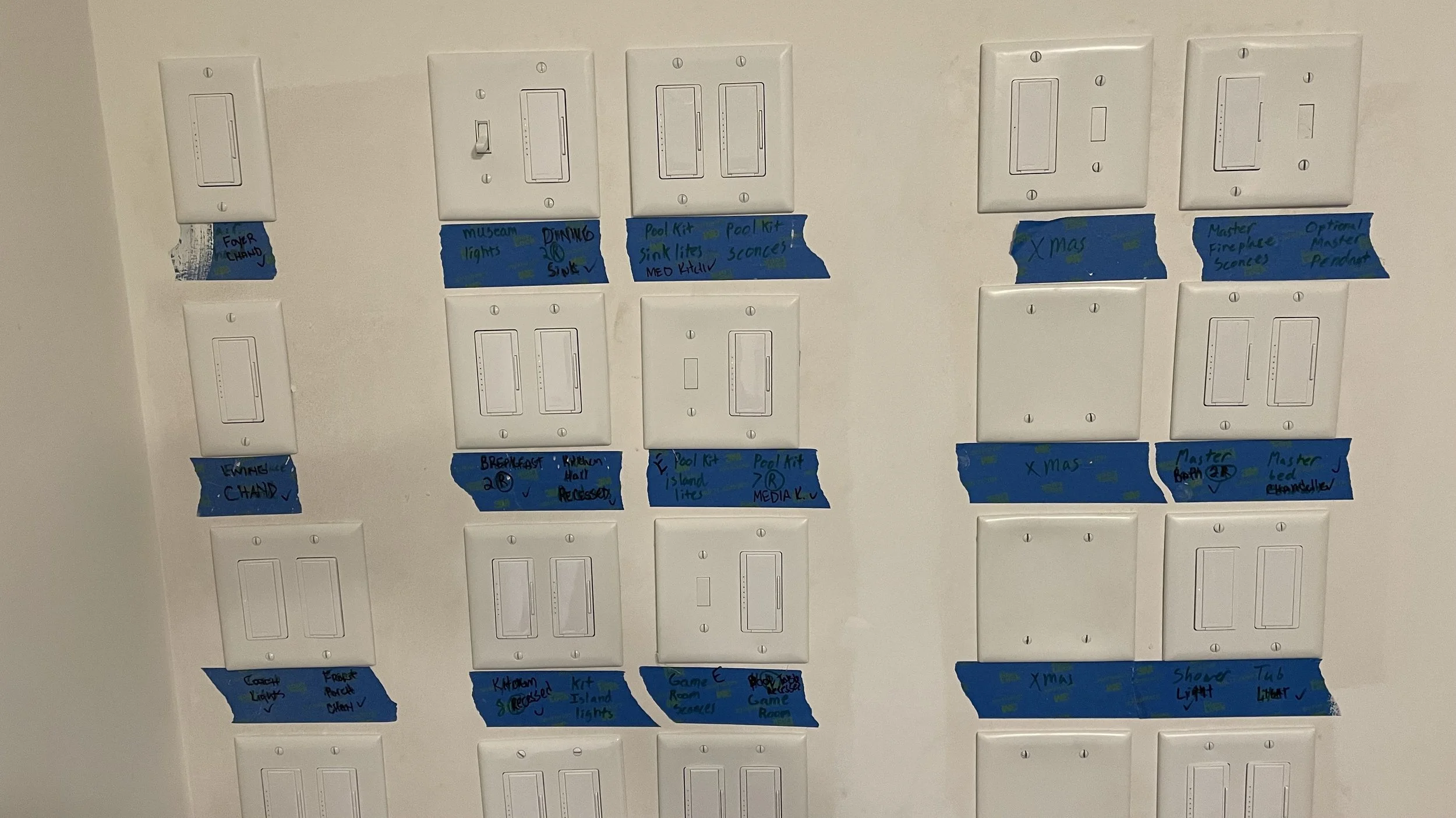 A wall with multiple Lutron Radio RA3 light switch panels, each with handwritten labels on blue painter's tape indicating different room lighting and scene settings.