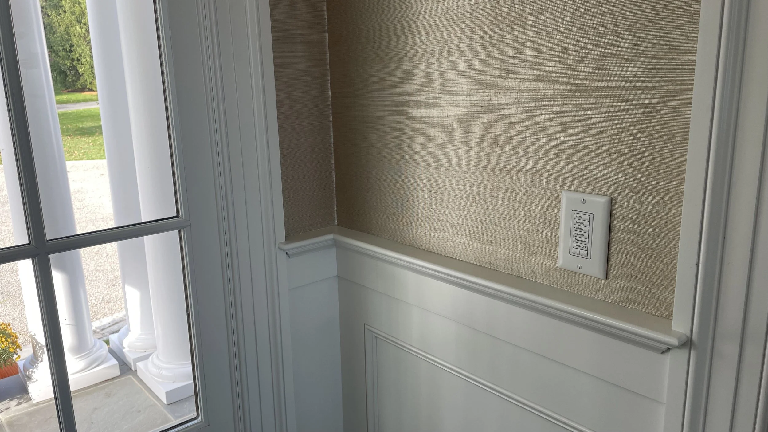 Interior view of a wall with a beige textured wallpaper, a white wainscoting panel, and a white wall control switch panel. Outside view through a glass door shows white columns and a patch of green grass.