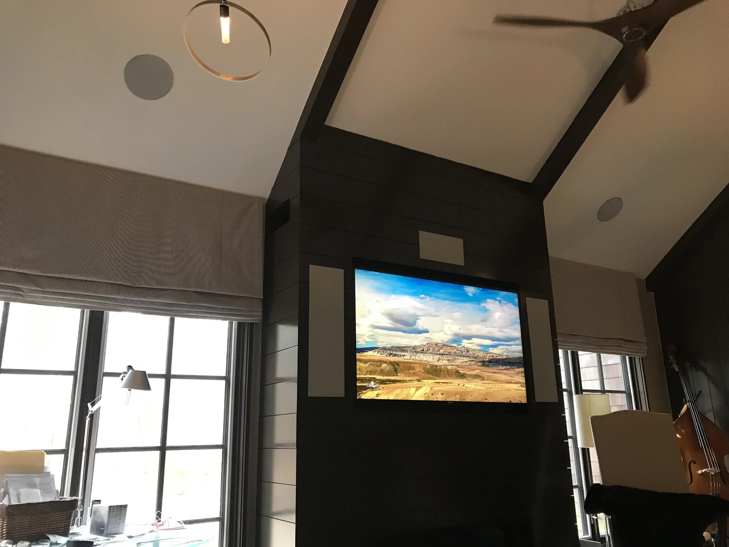 Living room with a television mounted on a dark wood-paneled wall, displaying a landscape with mountains and clouds, near two large windows with blinds, a cream-colored chair, and a side table with a lamp.