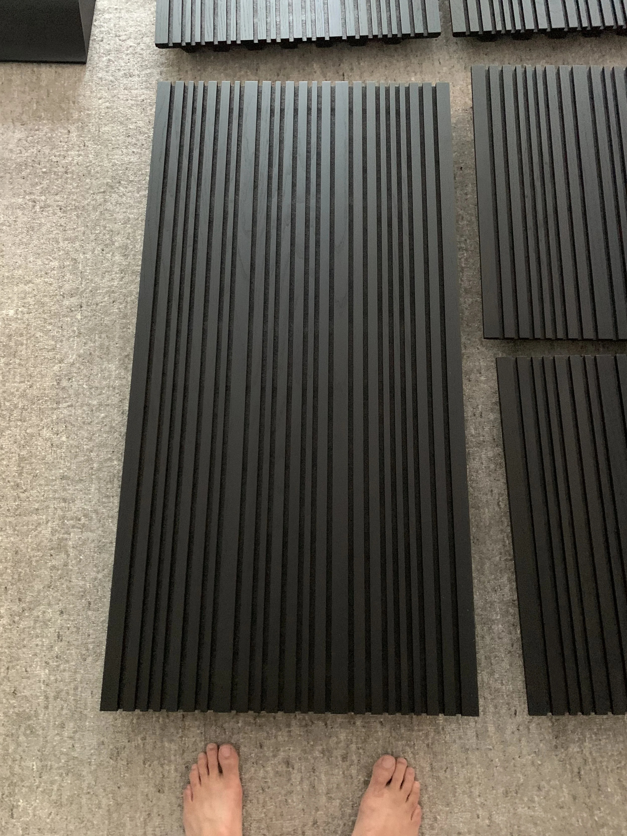 Someone's bare feet standing on a beige carpeted floor next to a set of black rubber or foam slats arranged on the floor.