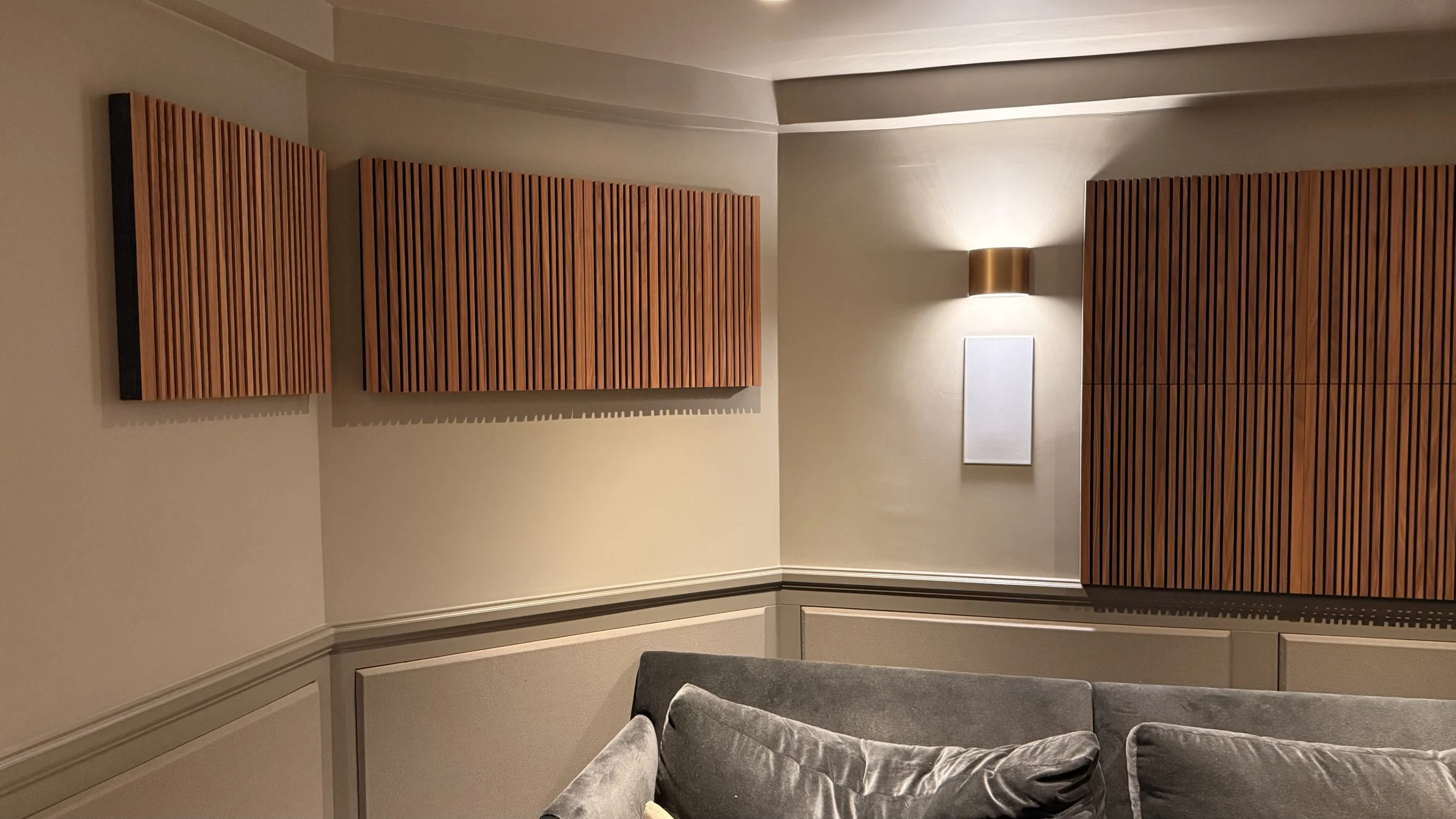 Interior of a room with beige walls, wooden acoustic panels on the wall, wall-mounted light fixture, rectangular white frame, and gray upholstery furniture.